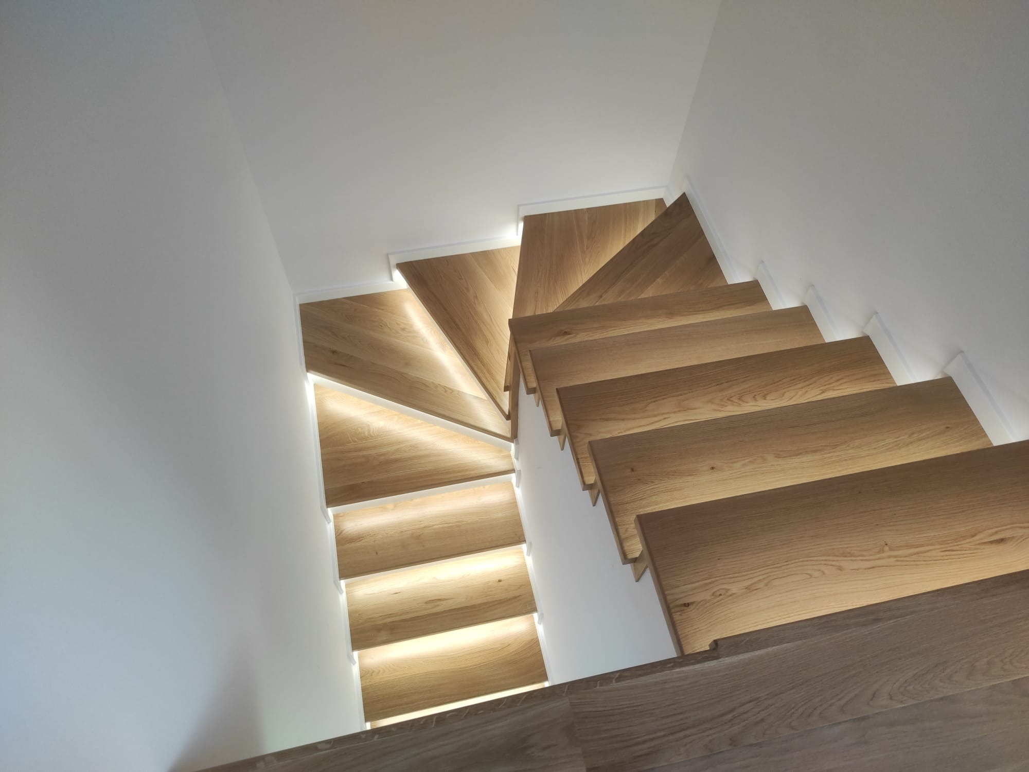 Drewniane schody z podświetleniem LED, widok z góry. Jasne drewno, białe ściany. Nowoczesny design, delikatne oświetlenie podkreśla strukturę drewna.