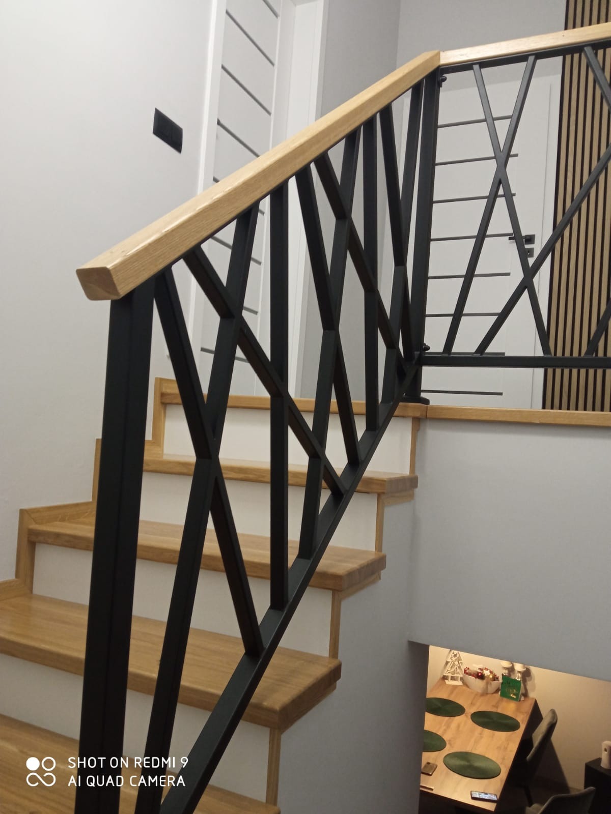 Nowoczesne schody drewniane z czarną, geometryczną balustradą. Jasne drewno stopni kontrastuje z ciemną konstrukcją. Widok z dołu, fragment jadalni w tle.