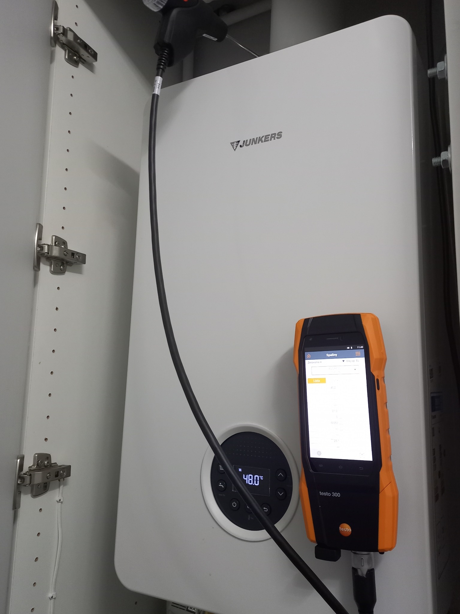 Biały piec gazowy Junkers w szafce, podłączony do analizatora spalin Testo 300. Widoczny kabel zasilający i wyświetlacz urządzenia pomiarowego.