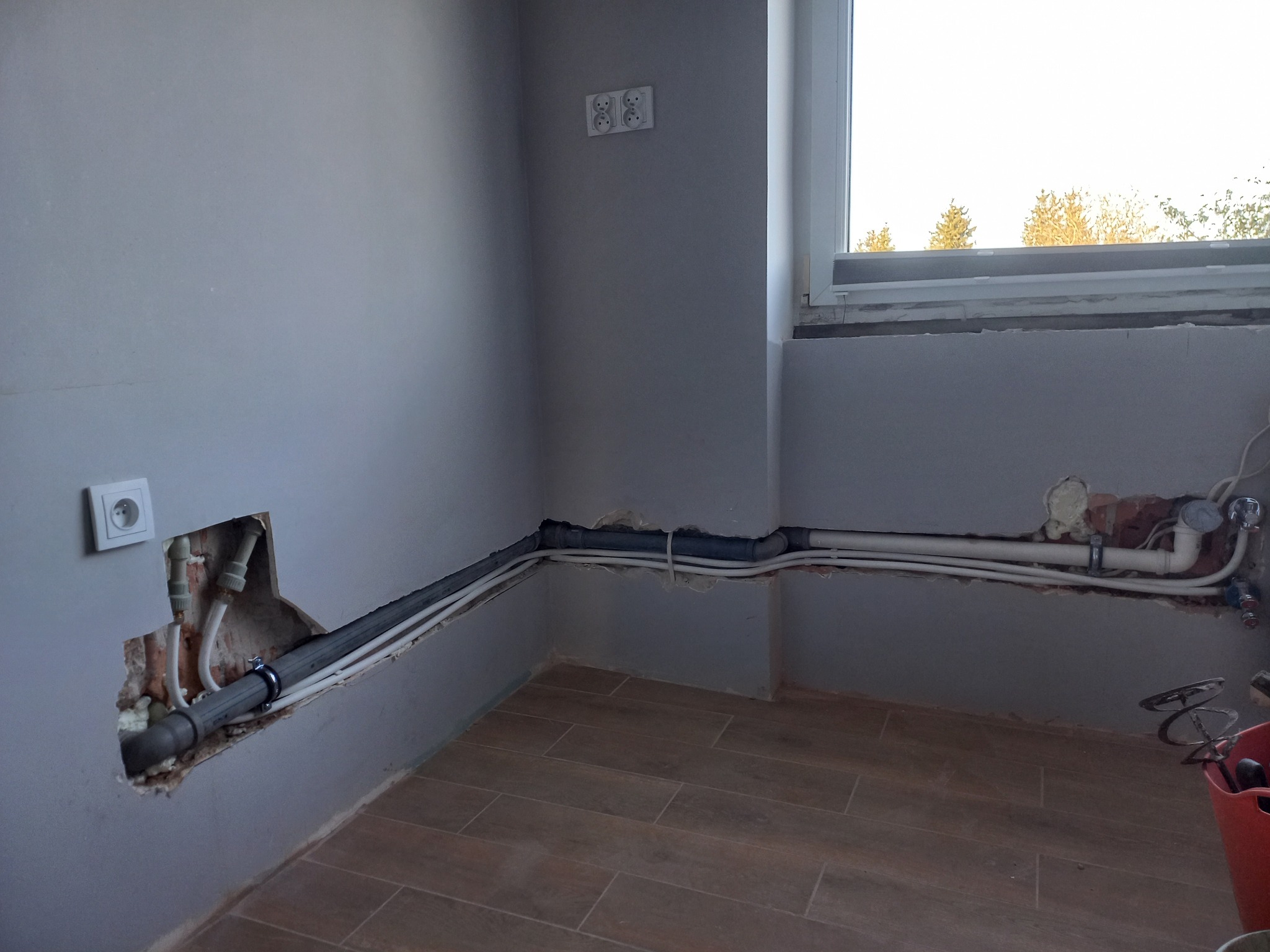 Szare wnętrze z widocznymi rurami instalacji wodnej i kanalizacyjnej, częściowo ukrytymi w ścianie. Widoczne gniazdka elektryczne i fragment okna.