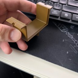 Dura3D - Beżowy, kątowy element z tworzywa, trzymany w dłoni na tle klawiatury i podkładki z grafiką techniczną. Widoczne warstwy sugerują wykonanie metodą addytywną.