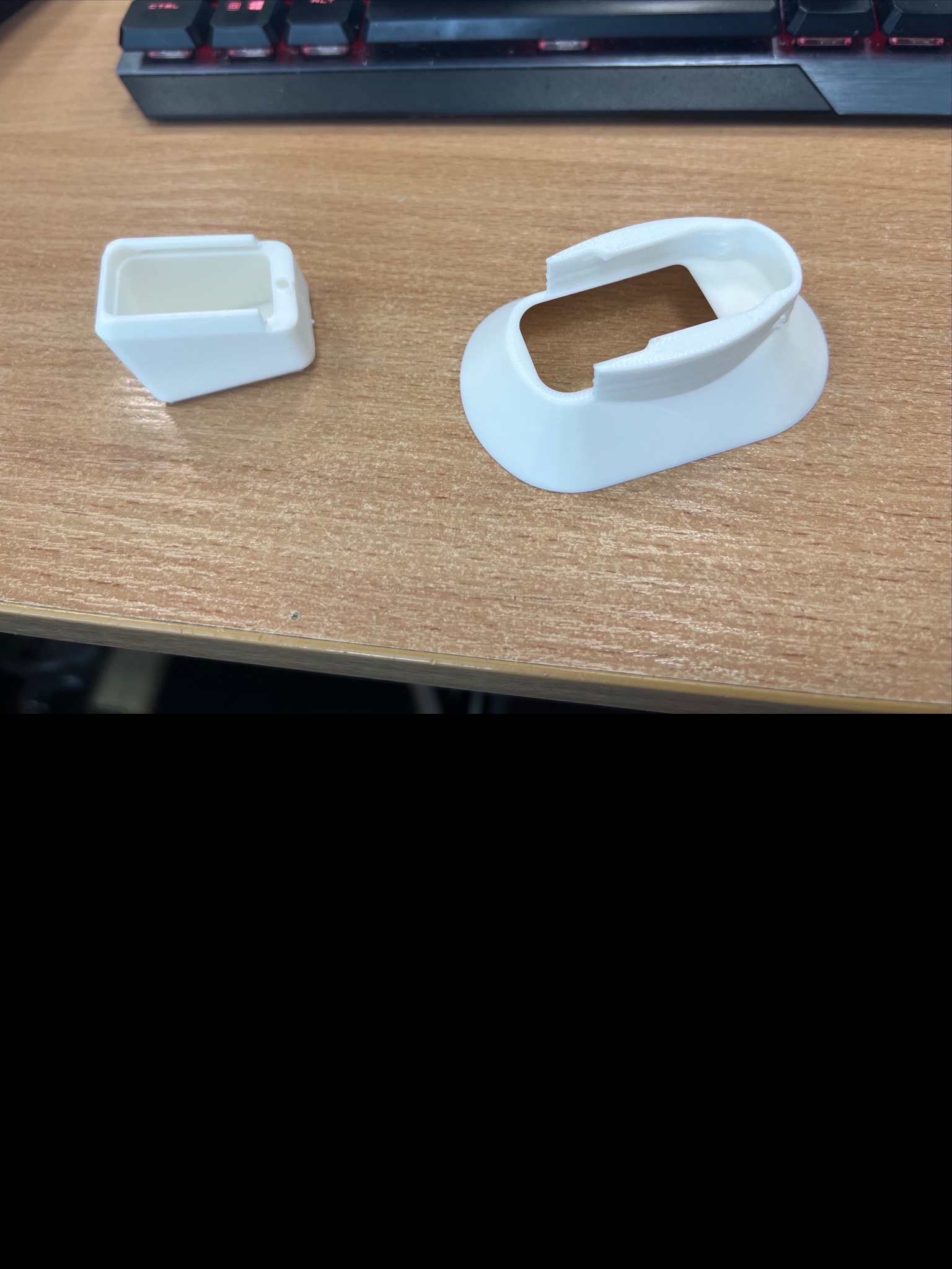 Dwa białe, plastikowe elementy wydrukowane w 3D leżą na drewnianym biurku obok klawiatury. Jeden element jest w kształcie sześcianu, drugi ma zaokrąglony kształt.