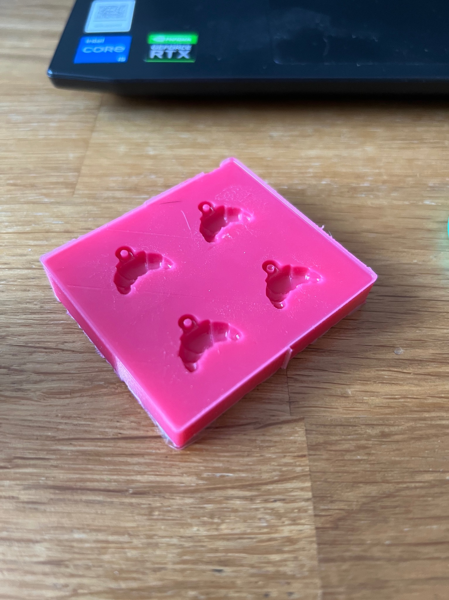 Różowa, silikonowa forma do odlewania z czterema wgłębieniami w kształcie rogala z oczkiem, leżąca na drewnianym blacie obok laptopa. Forma wykonana precyzyjnie.