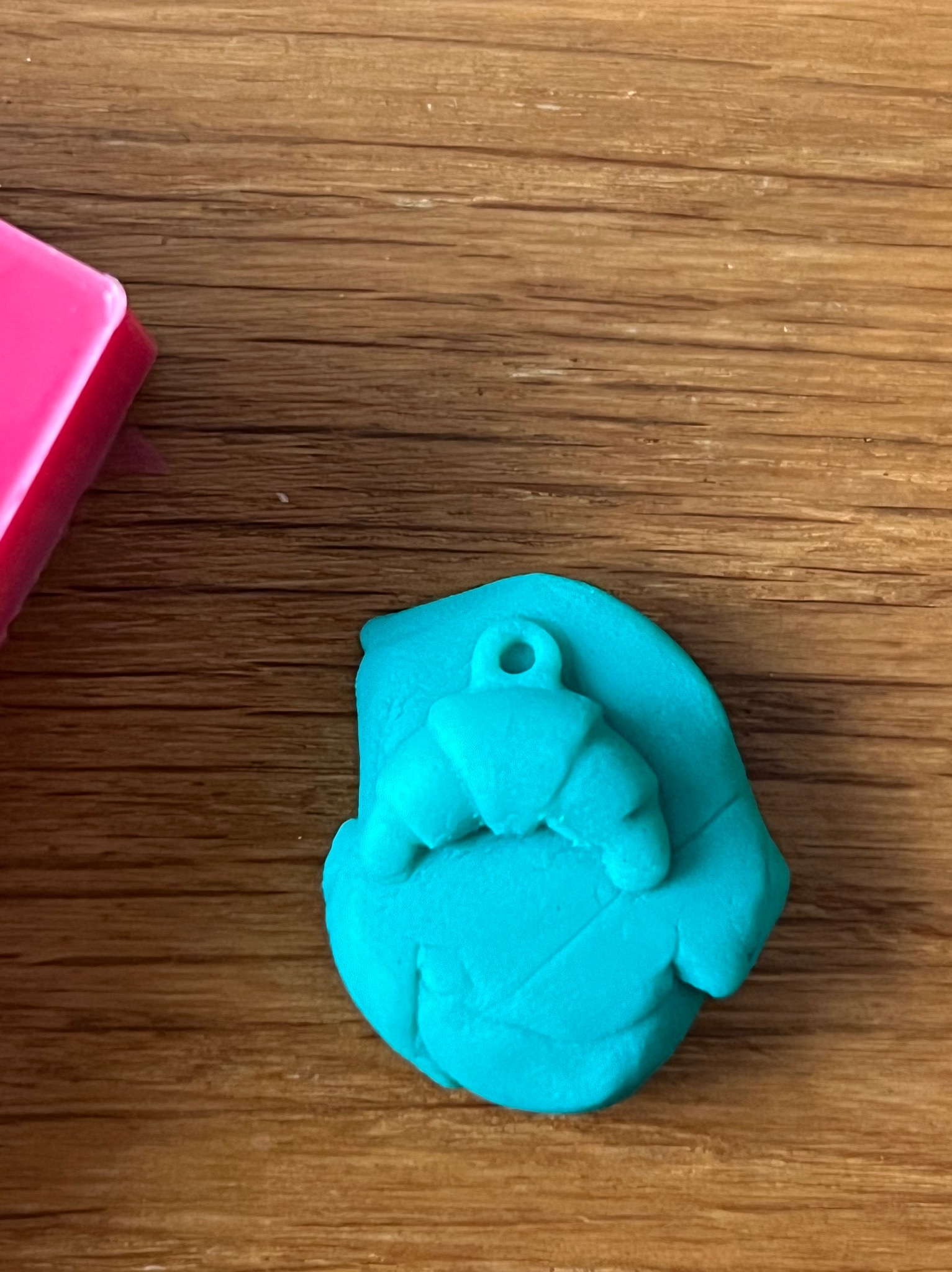 Turkusowy croissant z plasteliny z otworem na zawieszkę, leżący na drewnianym stole obok różowego pudełka. Modelowanie z plasteliny.