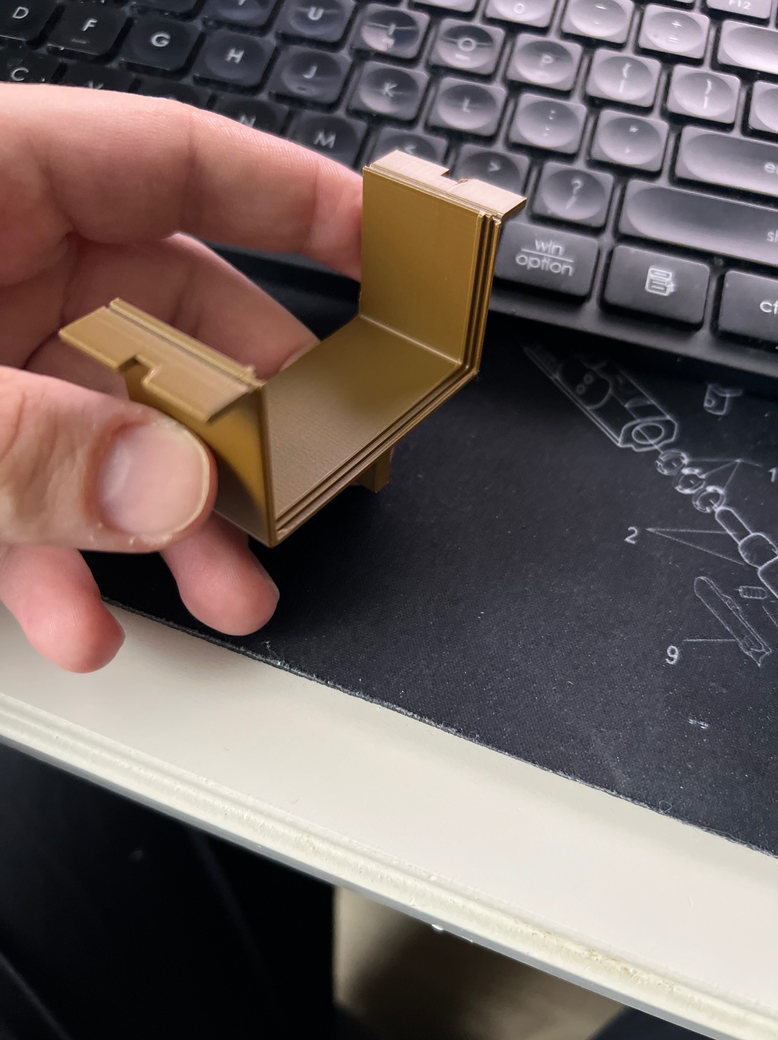 Beżowy, kątowy element z tworzywa, trzymany w dłoni na tle klawiatury i podkładki z grafiką techniczną. Widoczne warstwy sugerują wykonanie metodą addytywną.