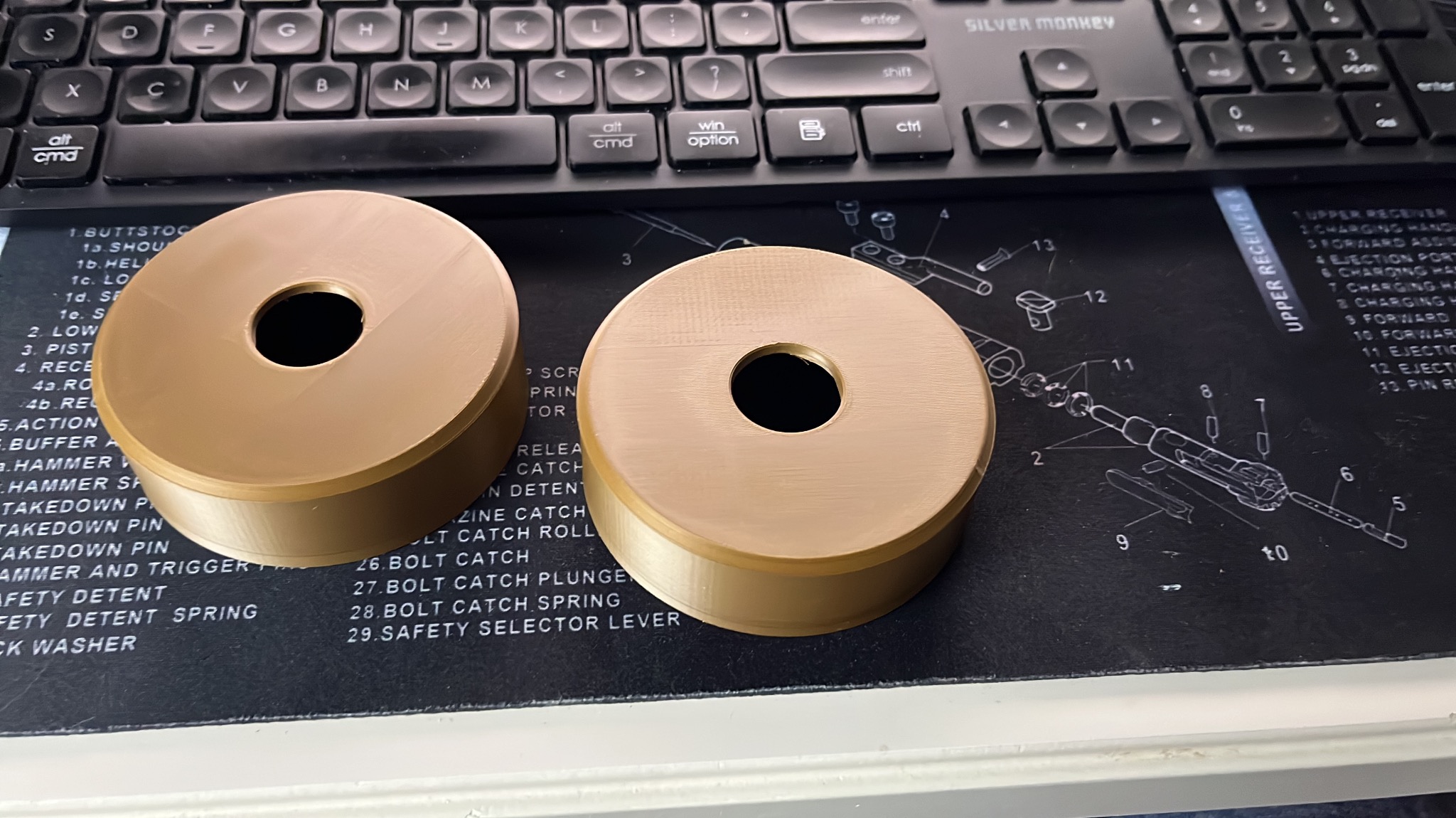 Dwa identyczne, beżowe, cylindryczne elementy z otworem, prawdopodobnie wykonane metodą addytywną, leżą na podkładce z schematem technicznym broni.