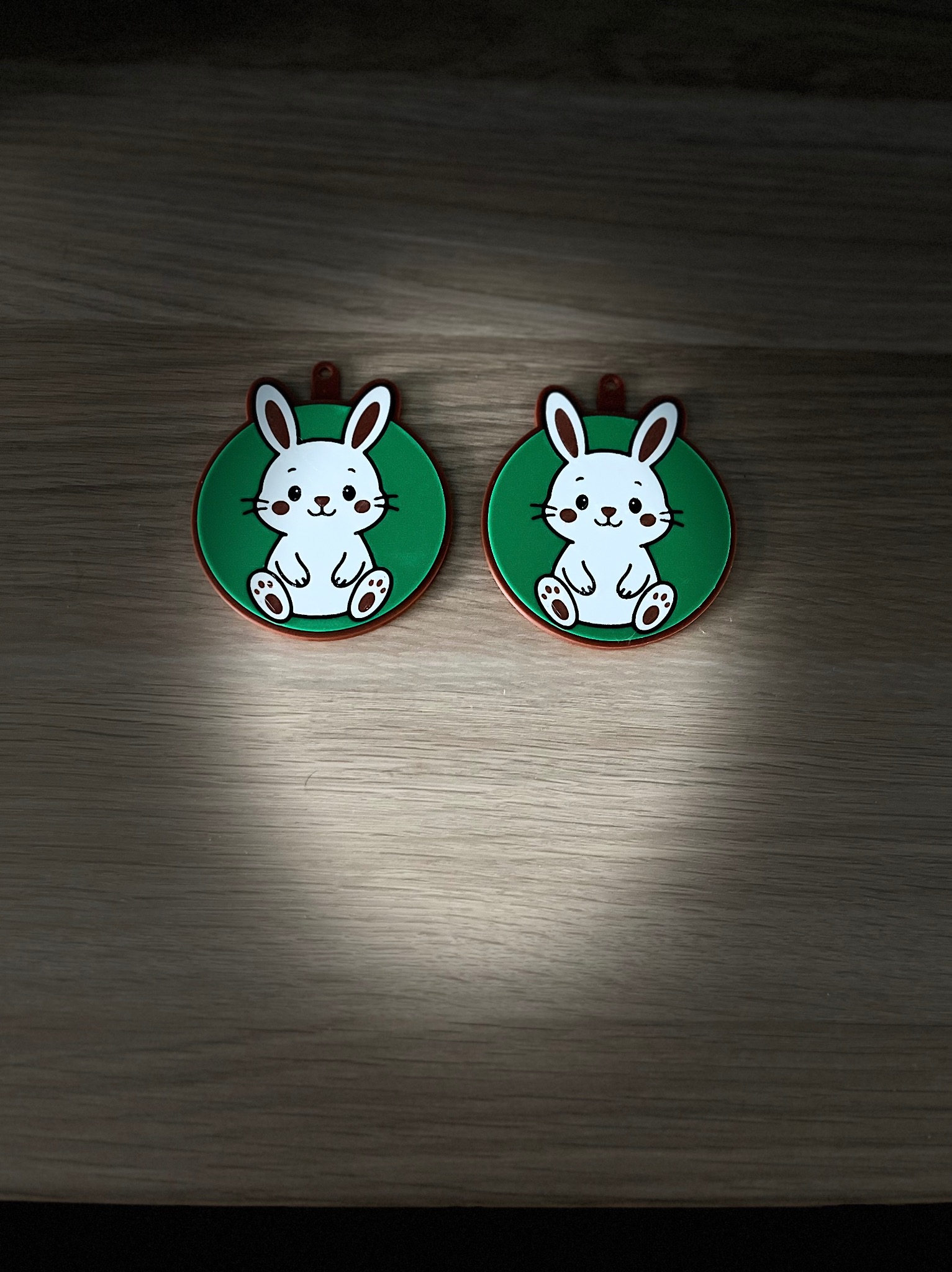 Dwa okrągłe breloczki z wizerunkiem białego królika na zielonym tle, leżące na drewnianej powierzchni w delikatnym oświetleniu.