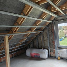 Eco Construction - Poddasze w trakcie adaptacji: widoczne metalowe profile konstrukcyjne, izolacja w folii, okno z widokiem na zewnątrz. Surowy stan, przygotowanie do dalszych prac wykończeniowych.