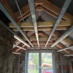 Eco Construction - Stelaż sufitu podwieszanego z profili metalowych w surowym wnętrzu poddasza z oknem, widok z dołu. Widoczne drewniane elementy konstrukcyjne dachu i fragmenty muru.