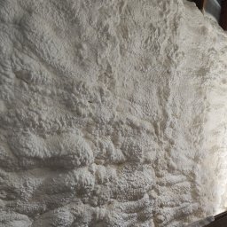 Eco Construction - Ściana pokryta grubą warstwą białej pianki izolacyjnej, widoczne fragmenty drewnianej konstrukcji i muru z białej cegły. Aplikacja pianki w trudno dostępnym miejscu.