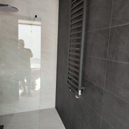 Eco Construction - Nowoczesna łazienka z szarymi płytkami, grzejnikiem drabinkowym i kabiną prysznicową ze szklaną ścianką. W lustrze odbija się osoba robiąca zdjęcie.