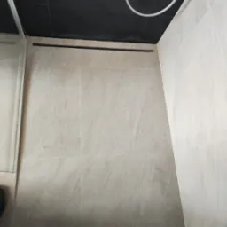 Wykończona kabina prysznicowa z odpływem liniowym, wyłożona beżowymi płytkami. Widoczny fragment szklanej ścianki i czarny sufit z elementami instalacji.