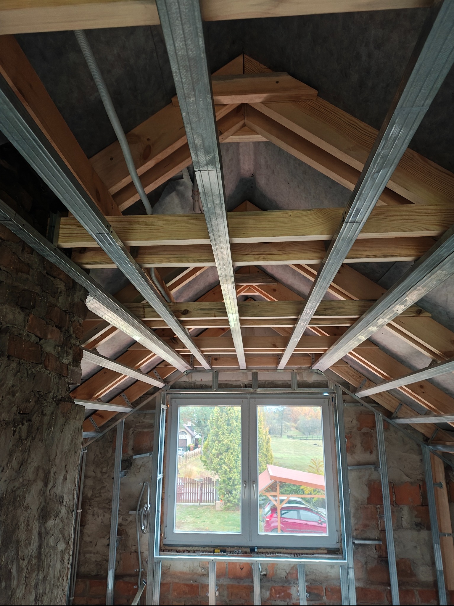 Stelaż sufitu podwieszanego z profili metalowych w surowym wnętrzu poddasza z oknem, widok z dołu. Widoczne drewniane elementy konstrukcyjne dachu i fragmenty muru.