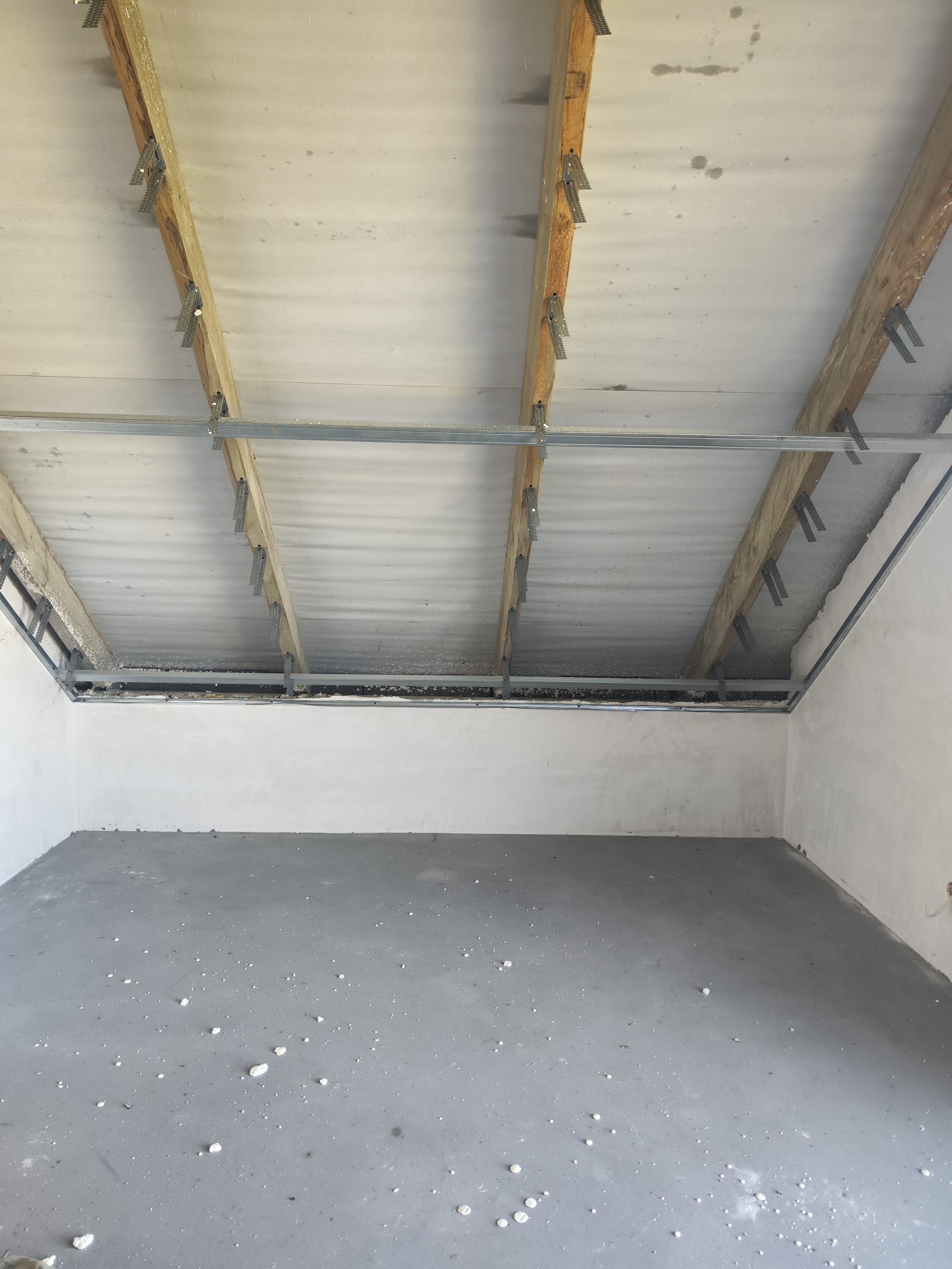 Surowe wnętrze z drewnianą konstrukcją sufitu i metalowymi profilami, przygotowane do dalszych prac wykończeniowych. Szara posadzka z resztkami materiałów budowlanych.