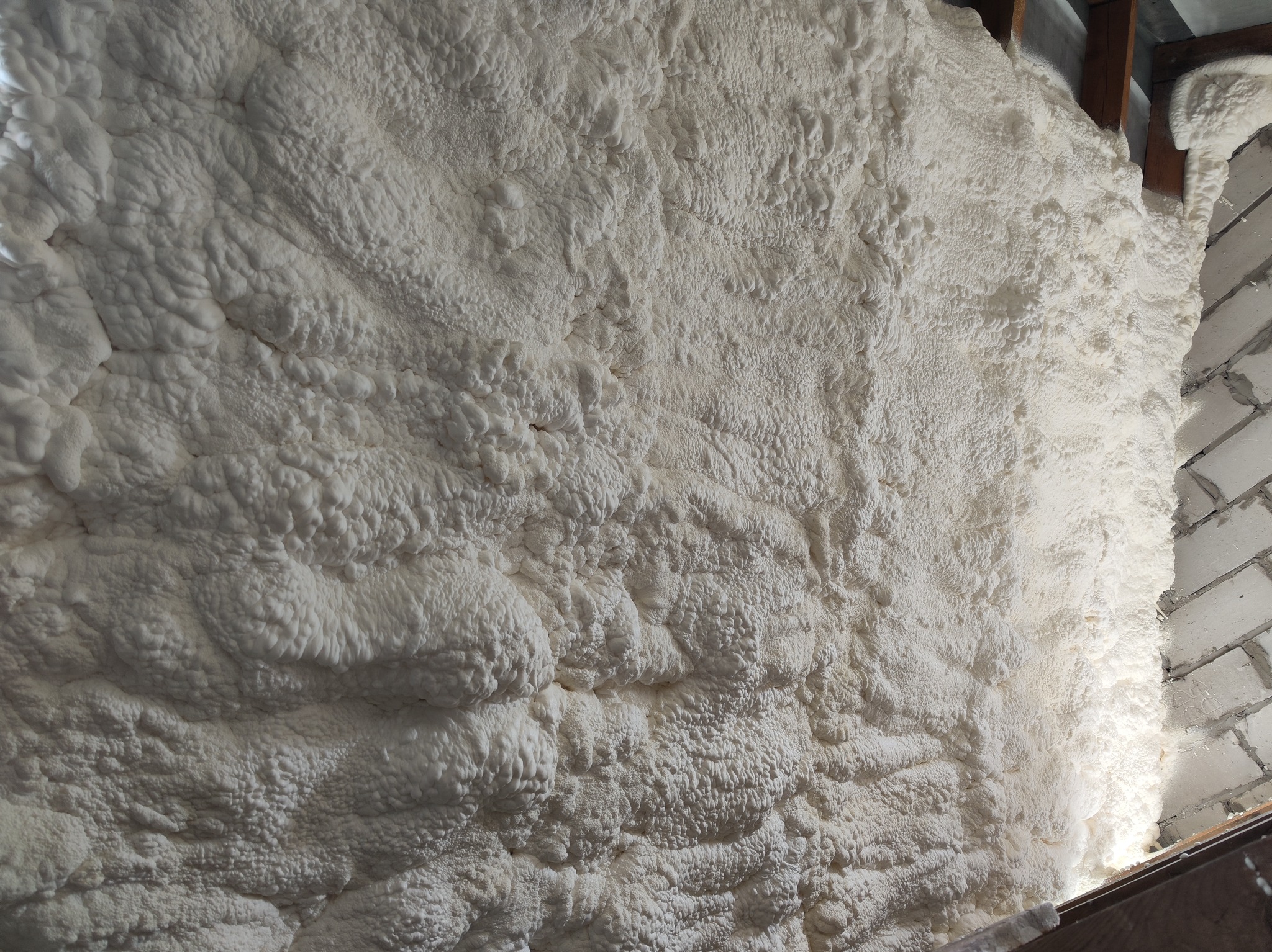 Ściana pokryta grubą warstwą białej pianki izolacyjnej, widoczne fragmenty drewnianej konstrukcji i muru z białej cegły. Aplikacja pianki w trudno dostępnym miejscu.