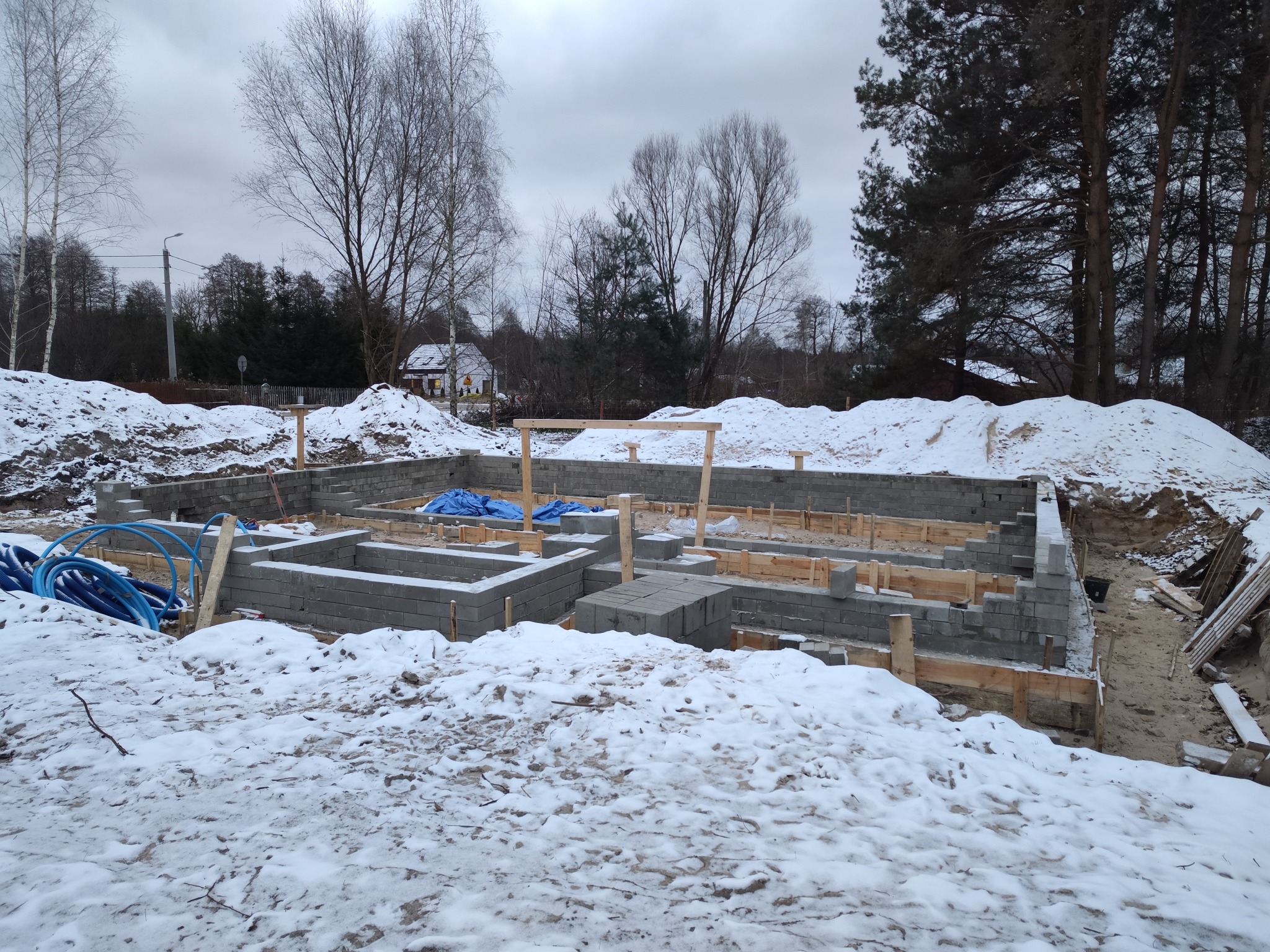 Zimowy widok na fundamenty domu w trakcie budowy, z widocznymi bloczkami betonowymi, drewnianym szalunkiem i niebieskimi rurami instalacyjnymi. Śnieg pokrywa teren.