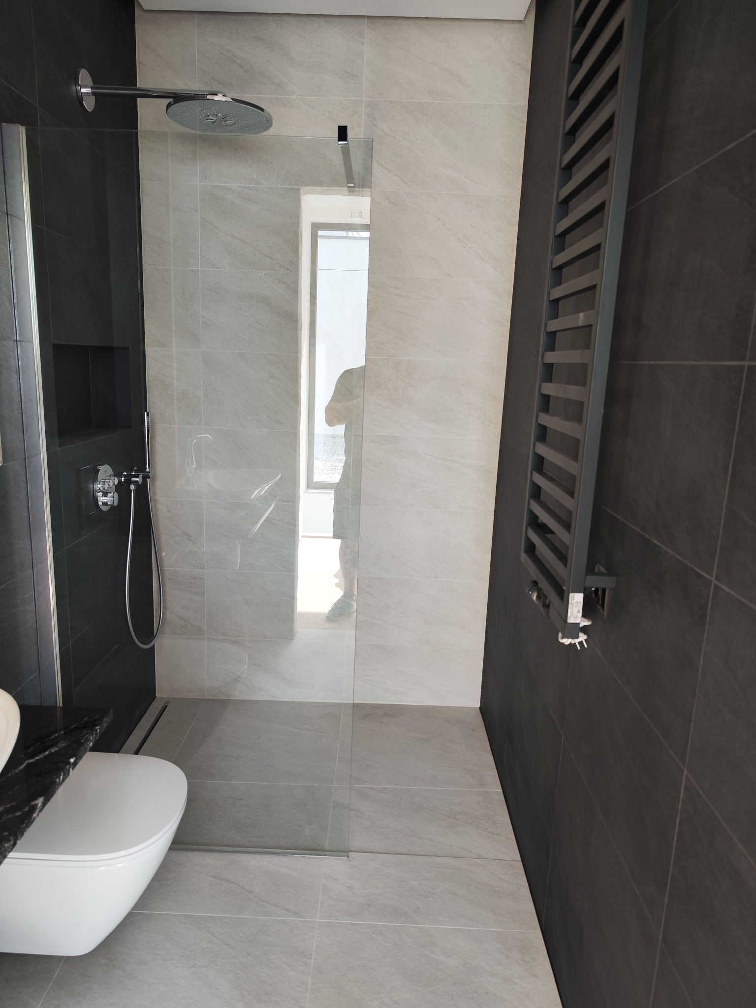 Nowoczesna łazienka z kabiną typu walk-in, grzejnikiem drabinkowym i podtynkową armaturą. Minimalistyczny design z szarymi i czarnymi płytkami.