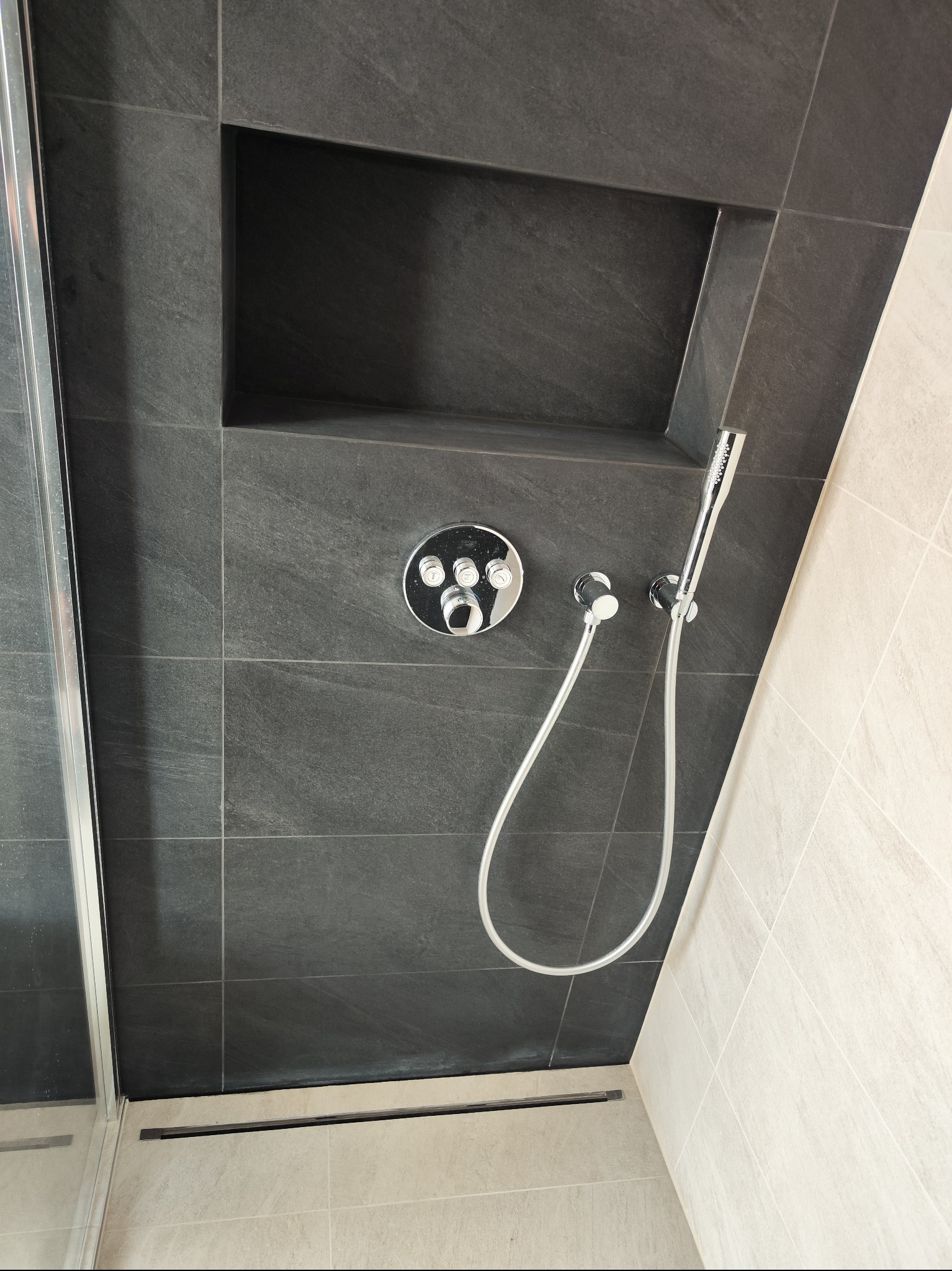 Nowoczesna łazienka z ciemnoszarą, kamienną ścianą prysznicową, wnęką i chromowaną armaturą. Widoczny fragment kabiny prysznicowej i jasne płytki na sąsiedniej ścianie.