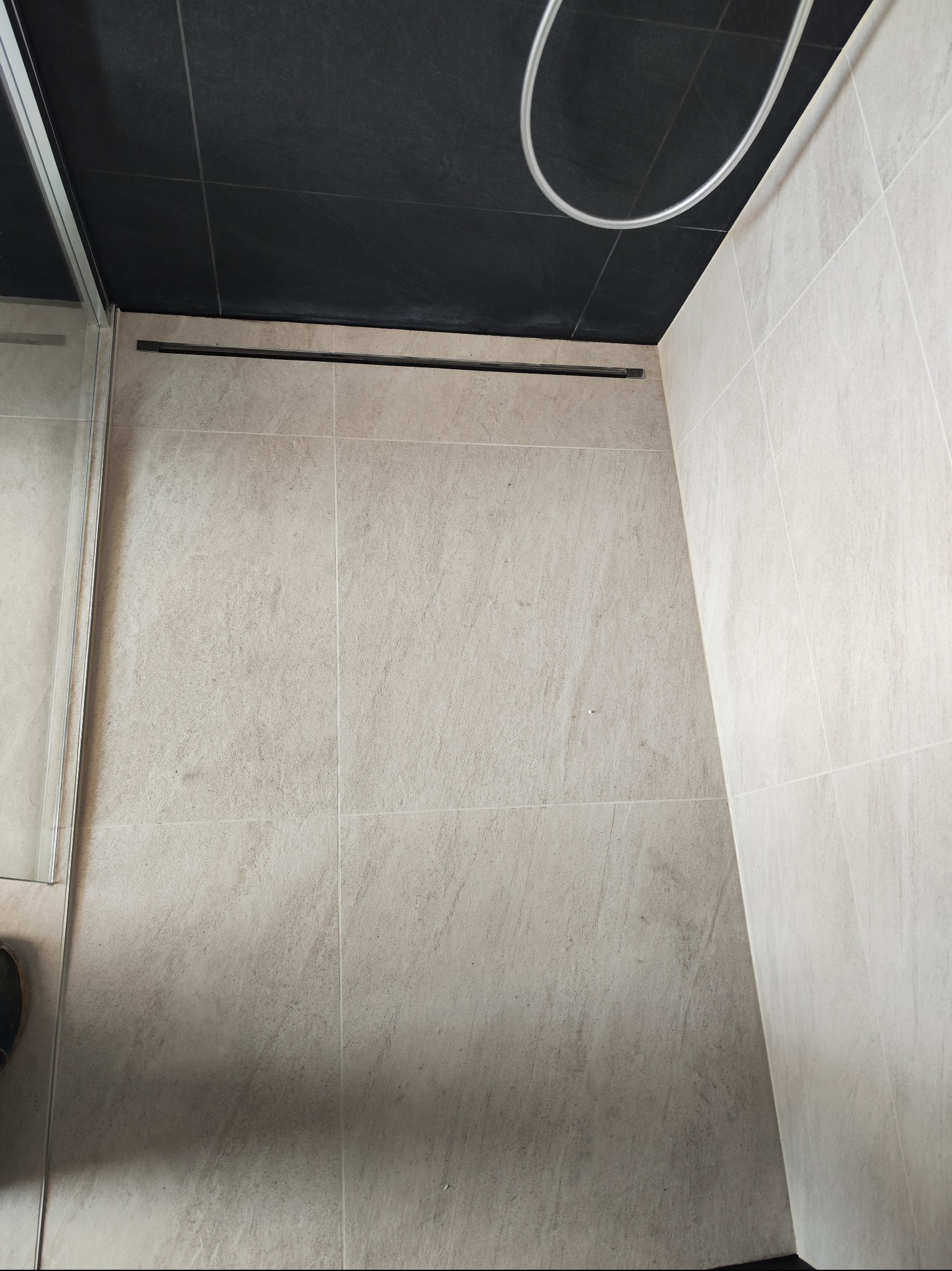 Wykończona kabina prysznicowa z odpływem liniowym, wyłożona beżowymi płytkami. Widoczny fragment szklanej ścianki i czarny sufit z elementami instalacji.