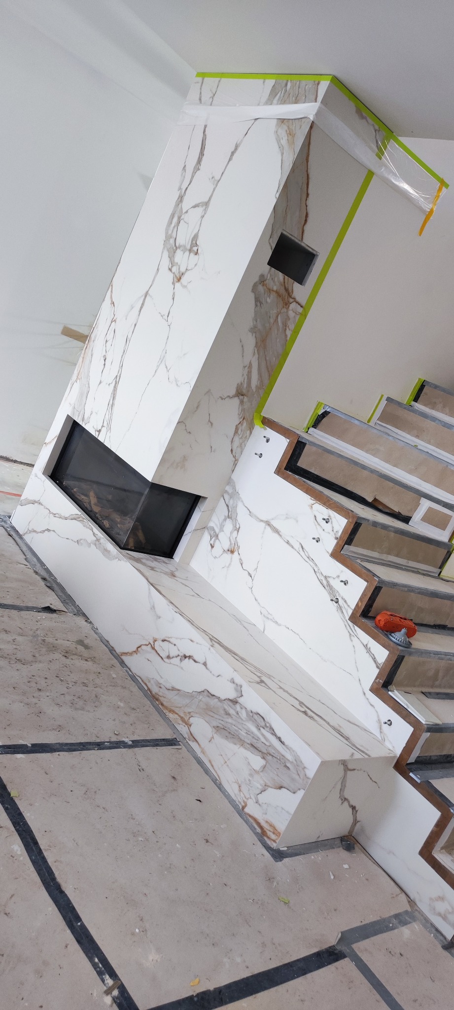 Obudowa kominka z płyt imitujących marmur, z widocznym paleniskiem i fragmentem schodów w trakcie wykończenia. Na schodach leży szlifierka. Widoczne zabezpieczenia taśmą.