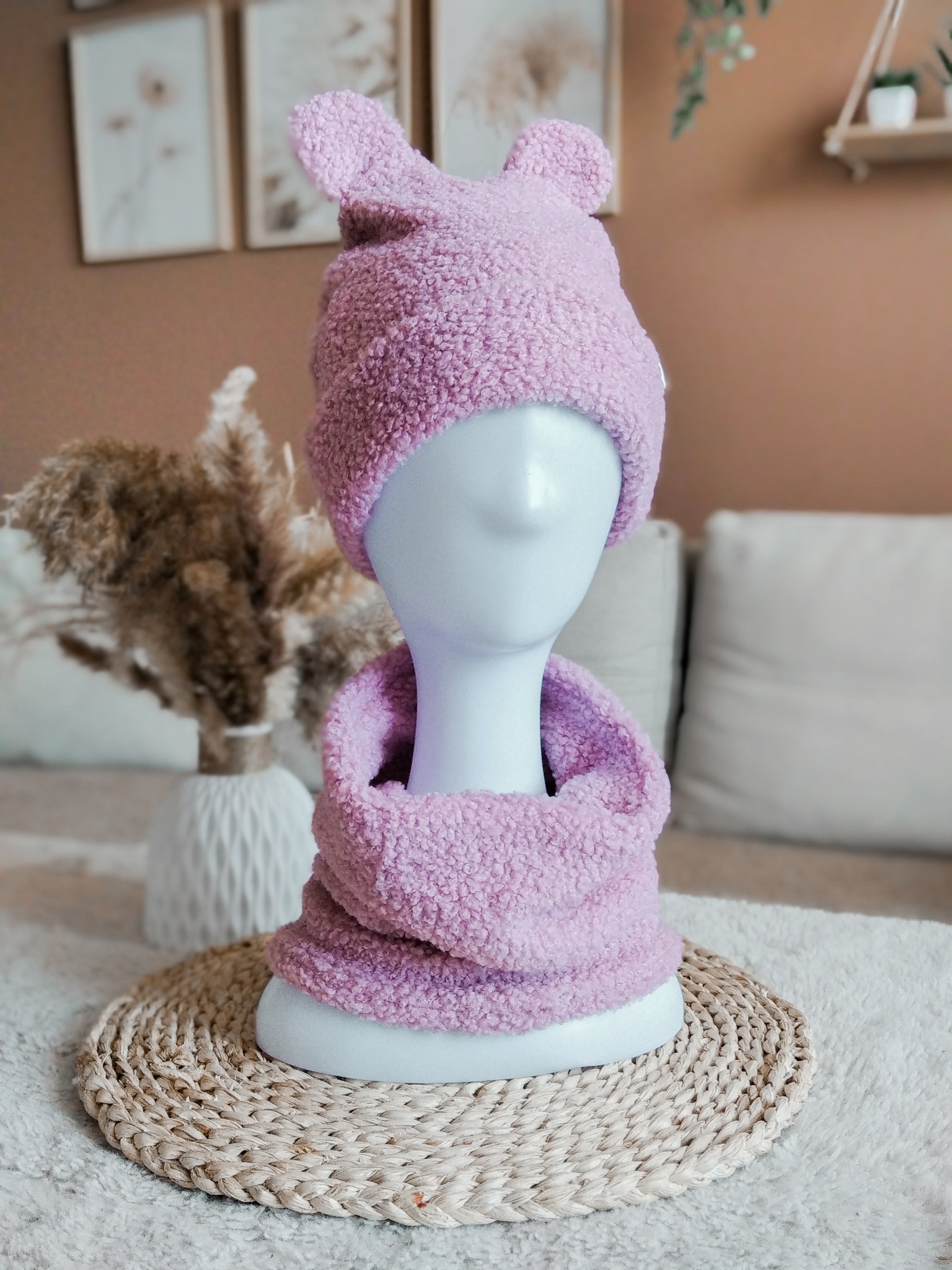 Różowy, pluszowy komplet: czapka z uszami i komin, na manekinie. W tle dekoracje w stylu boho i jasna sofa. Ujęcie w neutralnej tonacji.