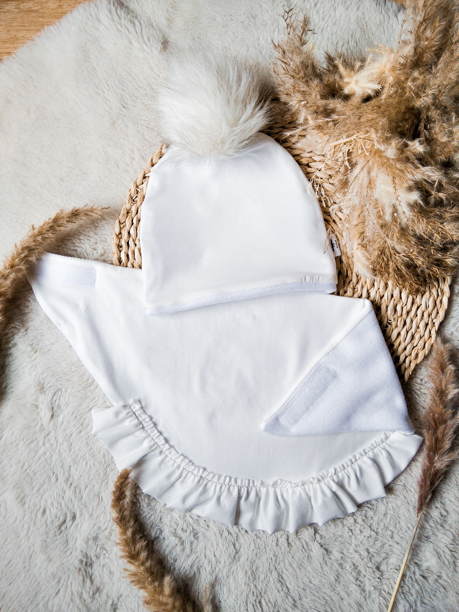 Biały komplet dziecięcy: czapka z pomponem i śliniak z falbanką, leżący na dekoracyjnym koszyku i puszystym dywanie, otoczony suszonymi trawami.