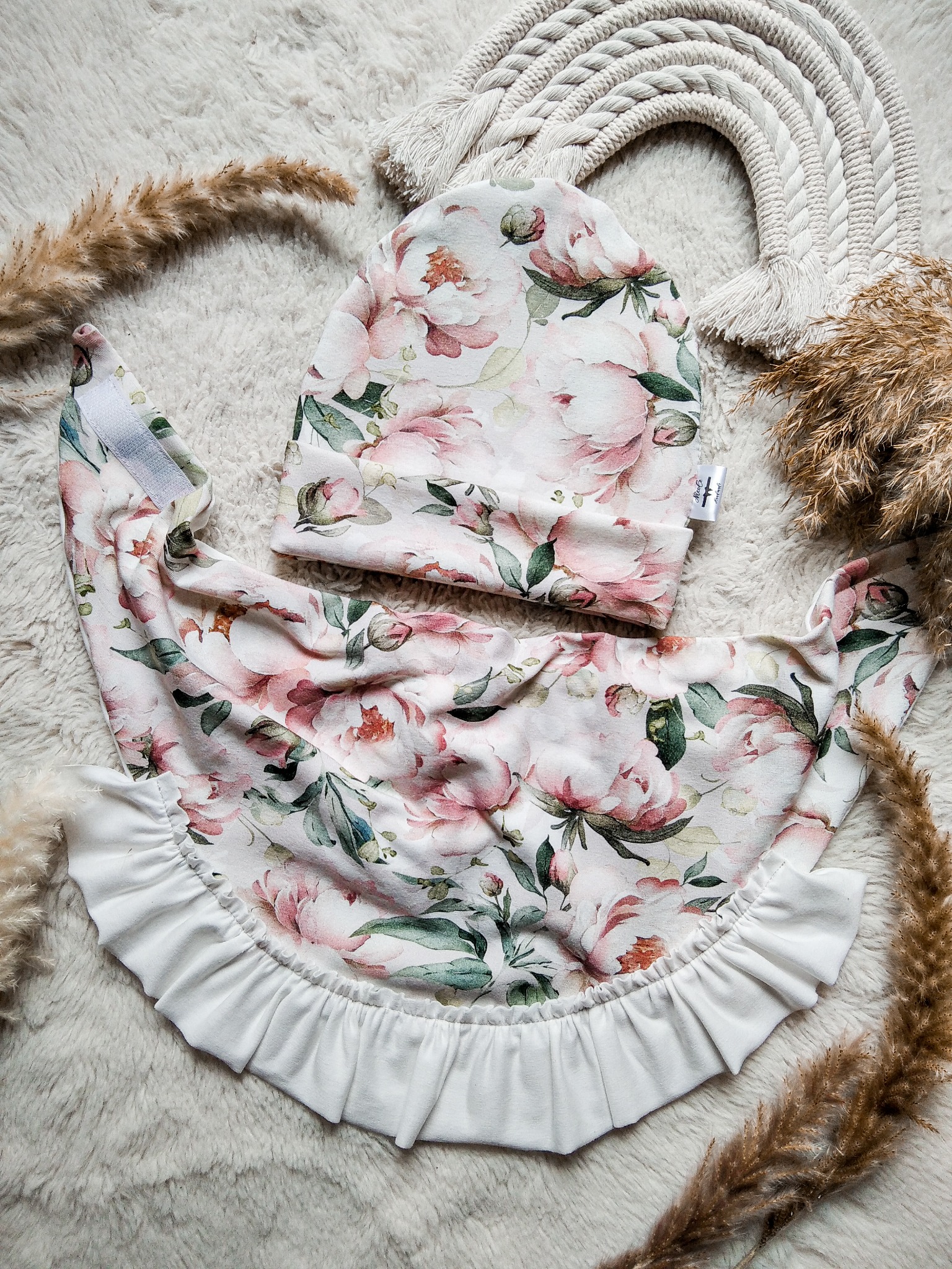 Ręcznie szyty komplet dziecięcy: czapka i śliniak z falbanką, w kwiatowy wzór, na puszystym tle z dekoracjami z suszonych traw i makramy.