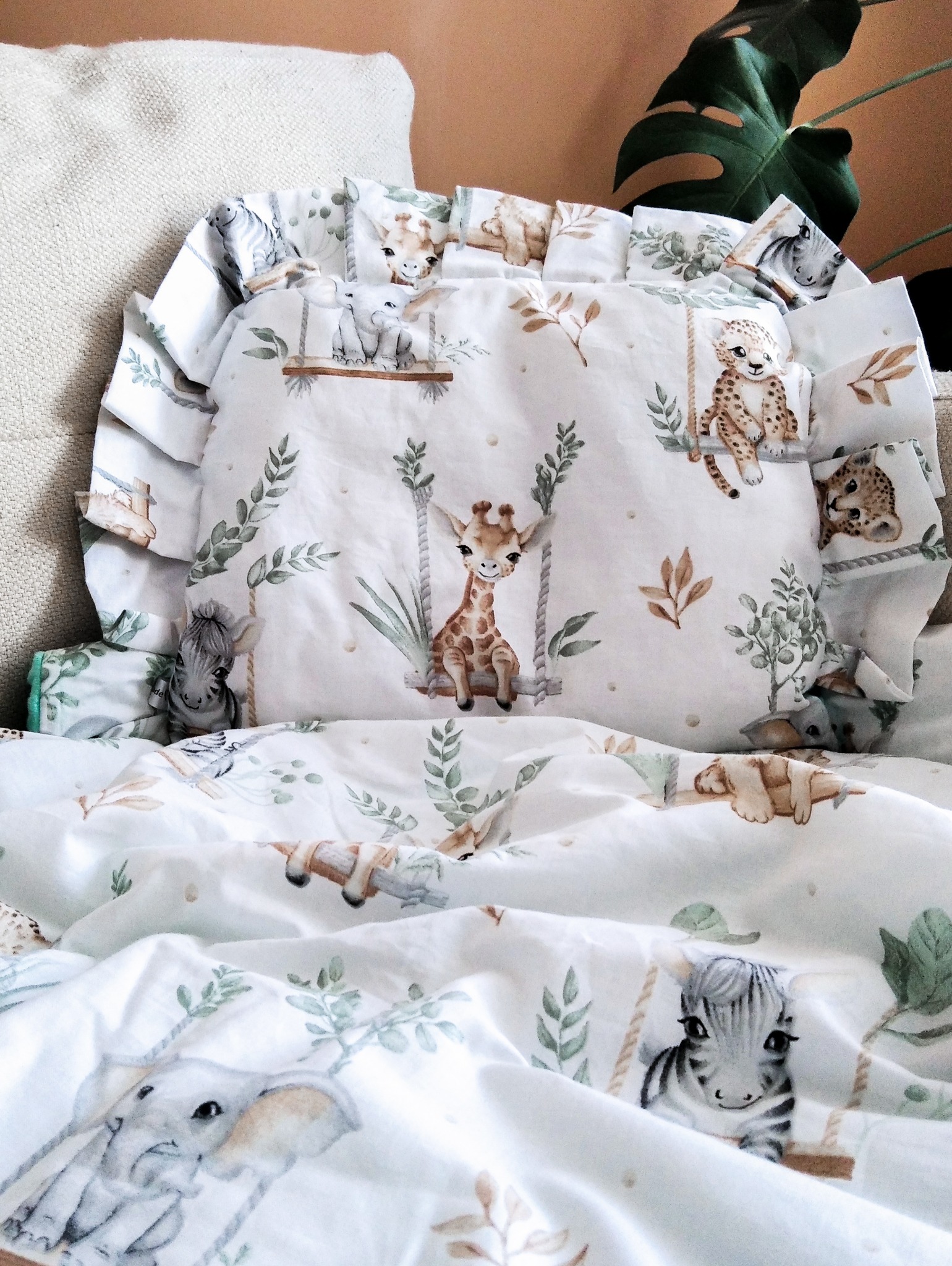 Dziecięca pościel z motywem zwierząt na huśtawkach: żyrafa, zebra, słoń, lampart. Biała tkanina w zielone liście, falbanki przy poduszce. Ujęcie z bliska.