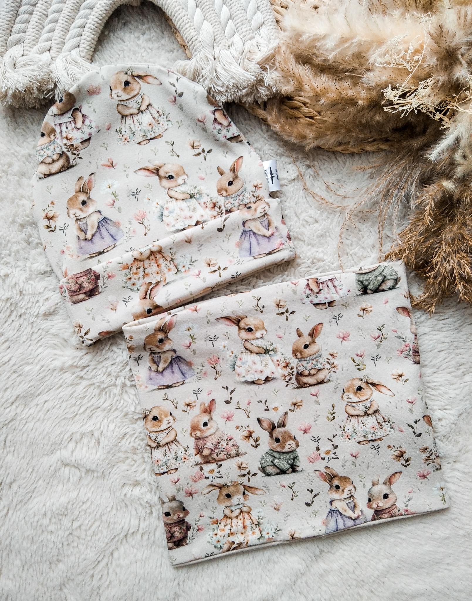 Dziecięca czapka i komin z motywem królików na miękkim, białym tle. Uroczy wzór, pastelowe kolory, idealne dla małych dzieci. Widoczna metka producenta.