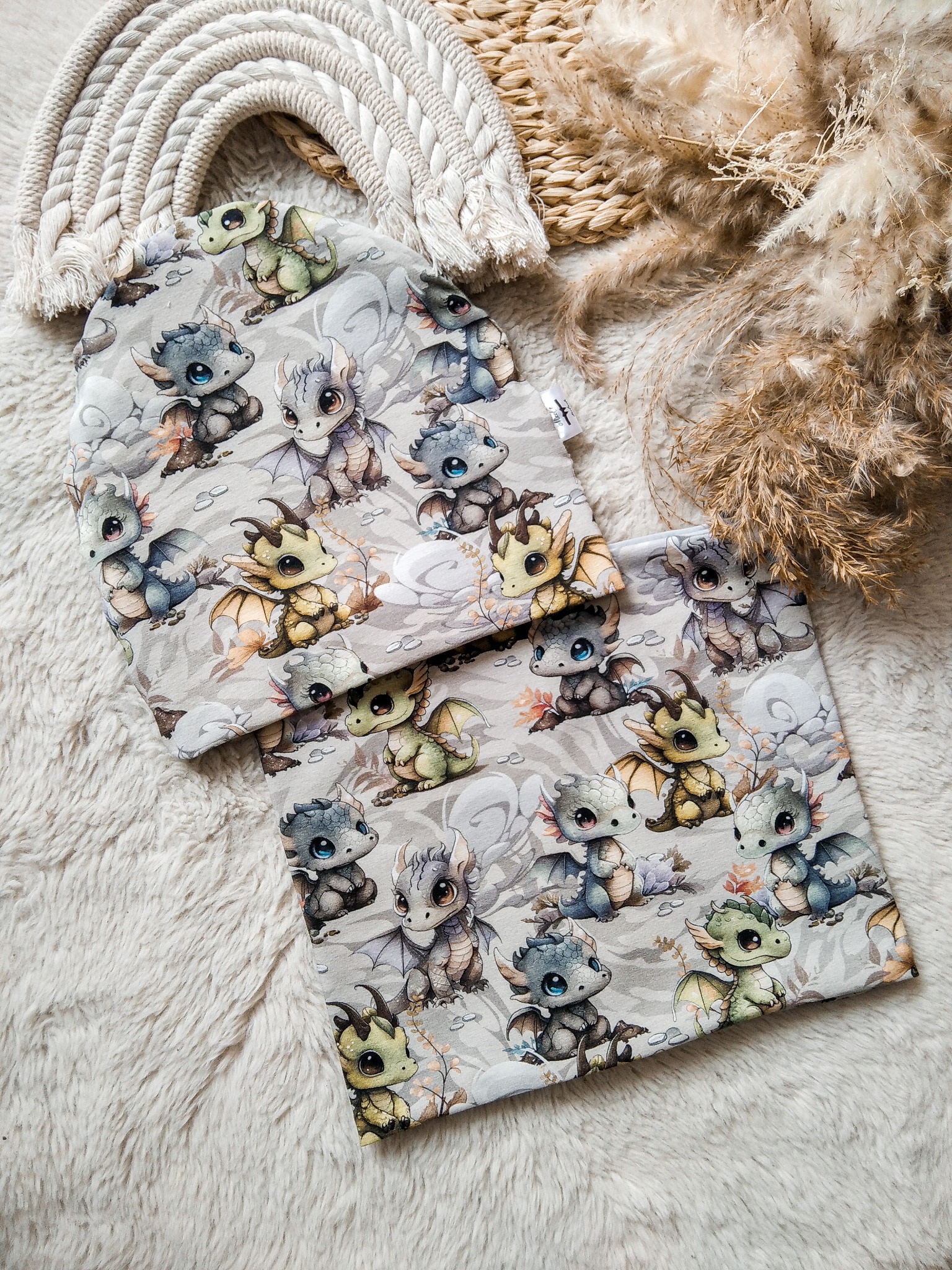 Dziecięca czapka i komin z motywem smoków na miękkim, białym tle, obok dekoracyjny łuk z sznurka i suszone trawy. Uroczy wzór i staranne wykonanie.