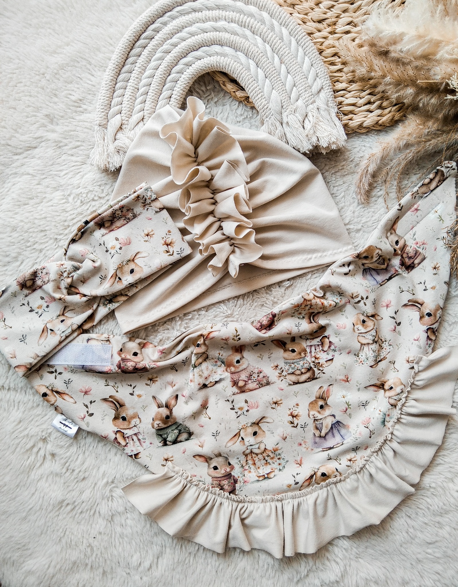Beżowy komplet dziecięcy: czapka z falbanką, opaska i śliniak z motywem królików na puszystym tle. Rękodzieło, pastelowa kolorystyka, delikatny wzór.