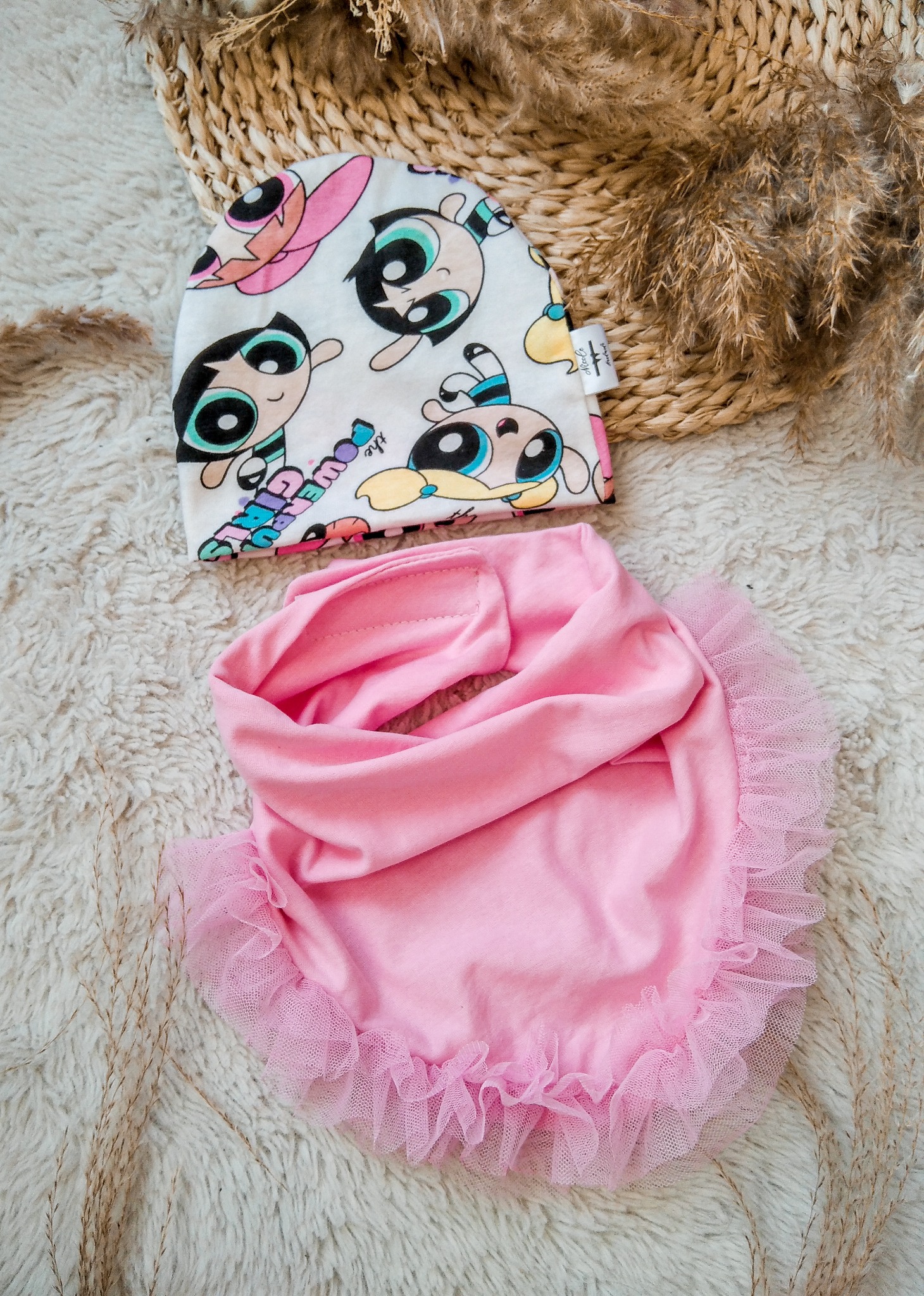 Ręcznie uszyty komplet dziecięcy: czapka z motywem bajkowym i różowy śliniak z falbanką, leżące na puszystym dywanie z elementami dekoracyjnymi.