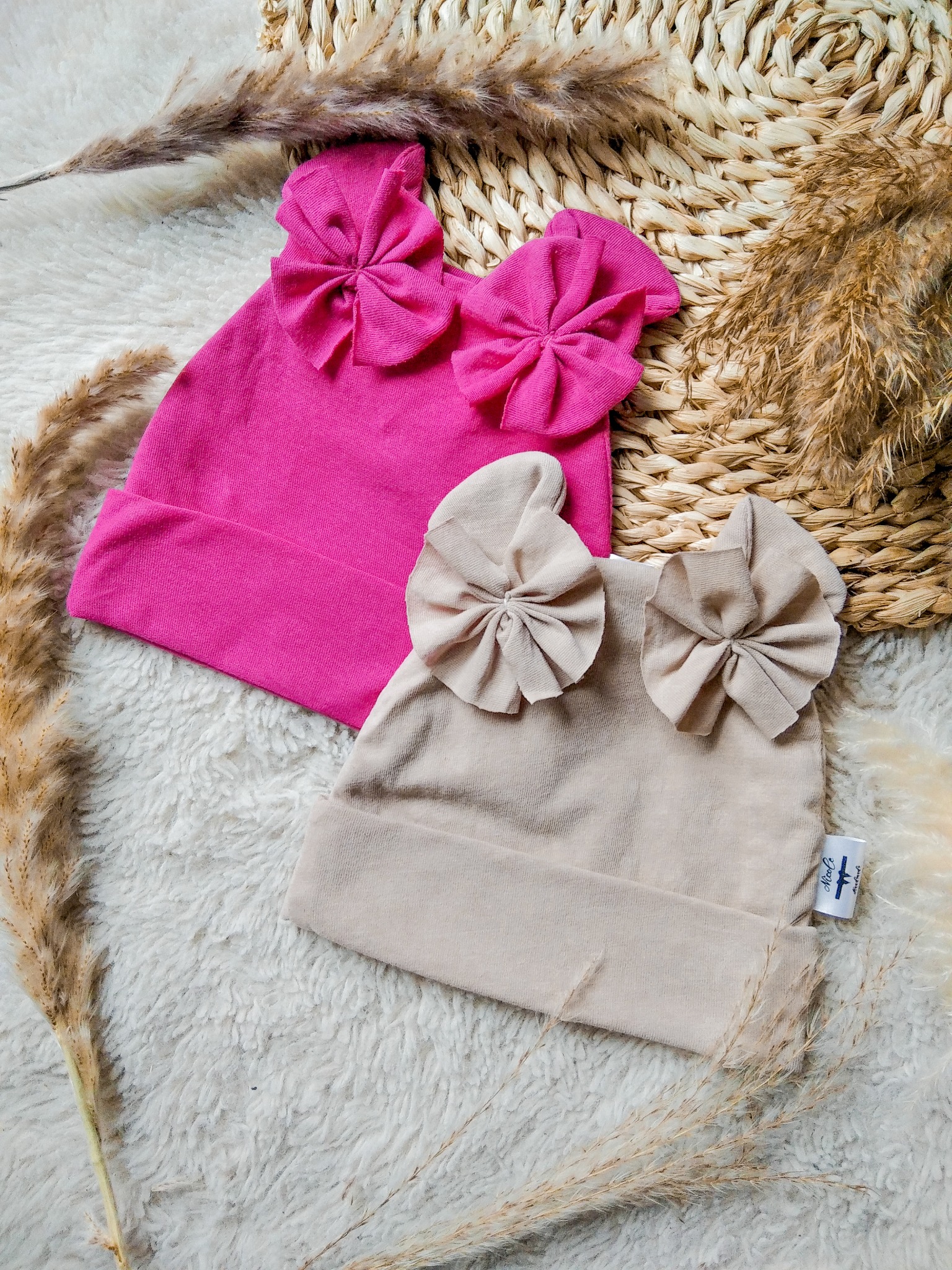 Różowa i beżowa czapka dziecięca z kokardami na puszystym tle, otoczone suszonymi trawami. Ujęcie z góry, prezentujące rękodzieło.