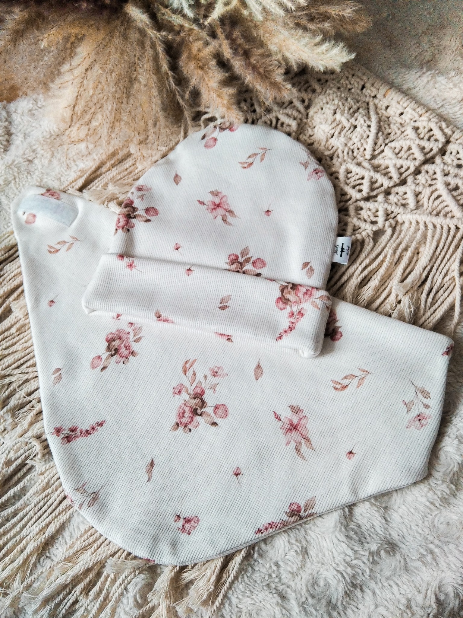 Komplet dziecięcy: czapka i śliniak z delikatnym, kwiatowym wzorem na kremowym tle, ułożone na beżowej makramie i puszystym dywanie. Widoczna metka.