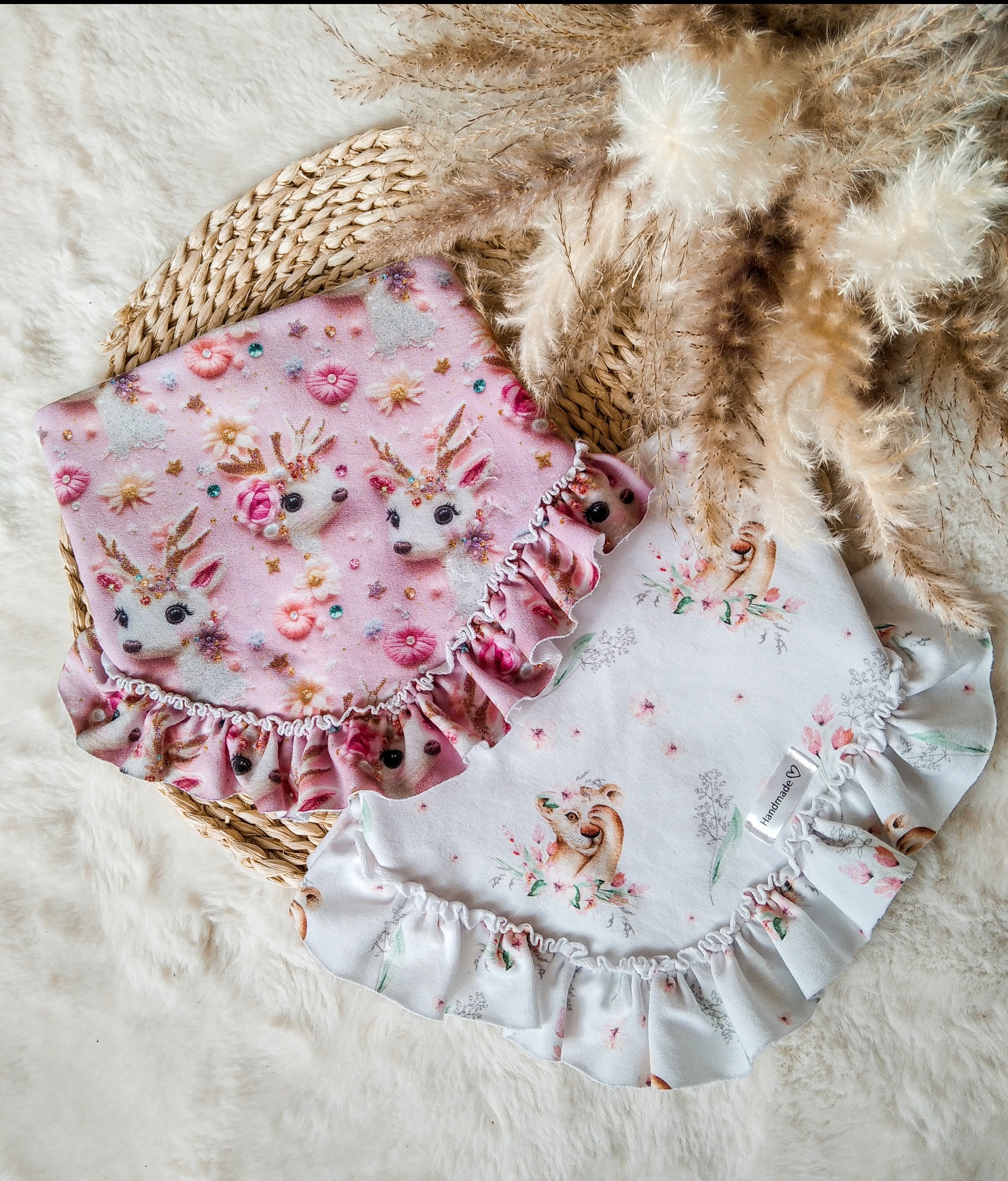 Dwa ręcznie szyte kocyki dziecięce z motywami zwierzęcymi i falbanką, leżące na wiklinowym podkładzie i puszystym futerku, otoczone suszonymi trawami.