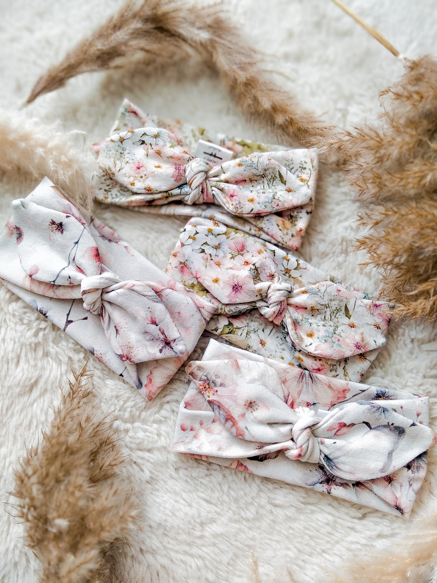 Trzy ręcznie szyte, kwieciste opaski dla dzieci z kokardami, ułożone na puszystym, białym tle z dekoracyjnymi trawami. Delikatne wzory kwiatowe, pastelowe kolory.