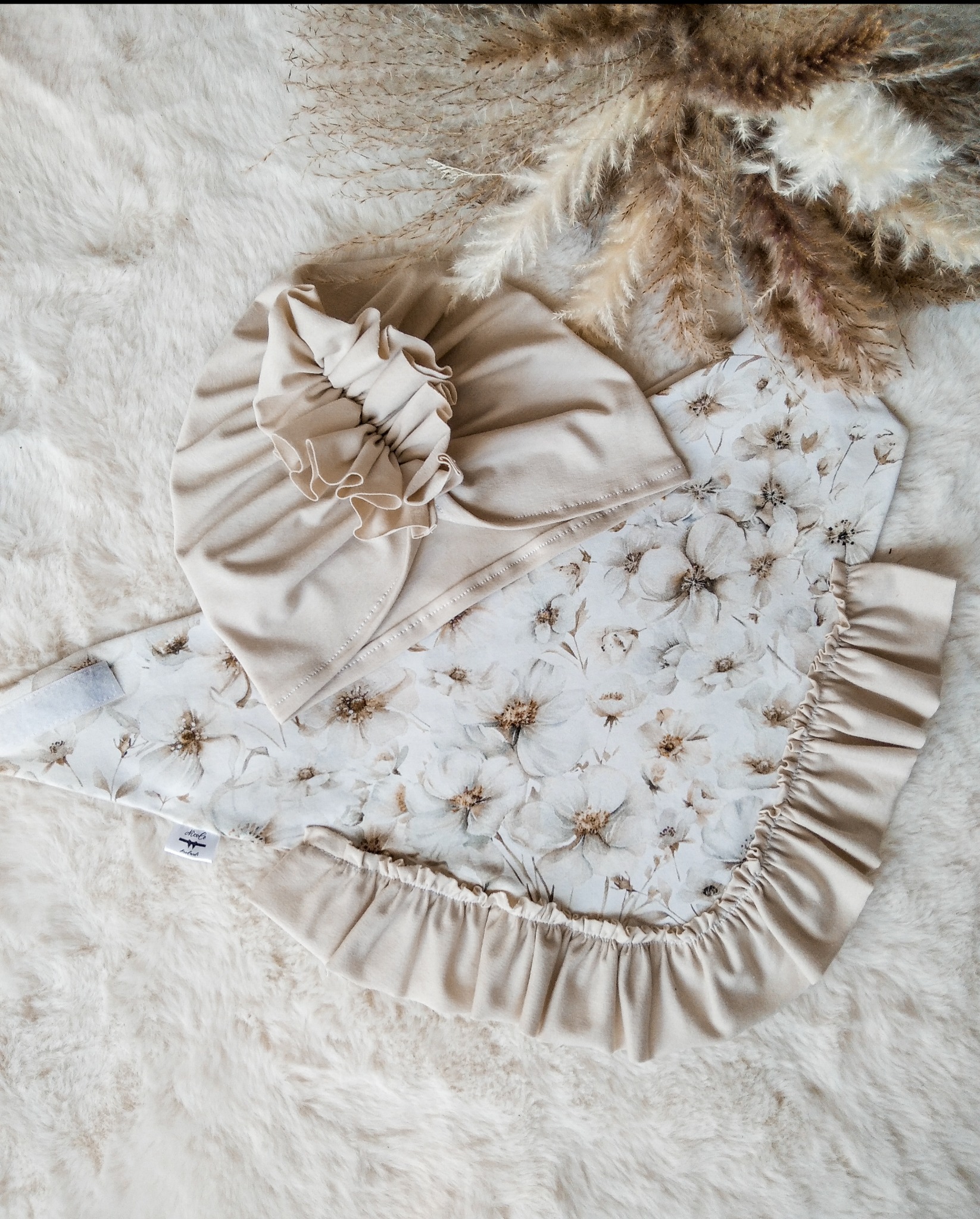 Beżowy komplet niemowlęcy: czapeczka z falbanką i kocyk w kwiaty, leżący na puszystym, kremowym futerku, obok dekoracyjne trawy pampasowe. Delikatna kolorystyka.