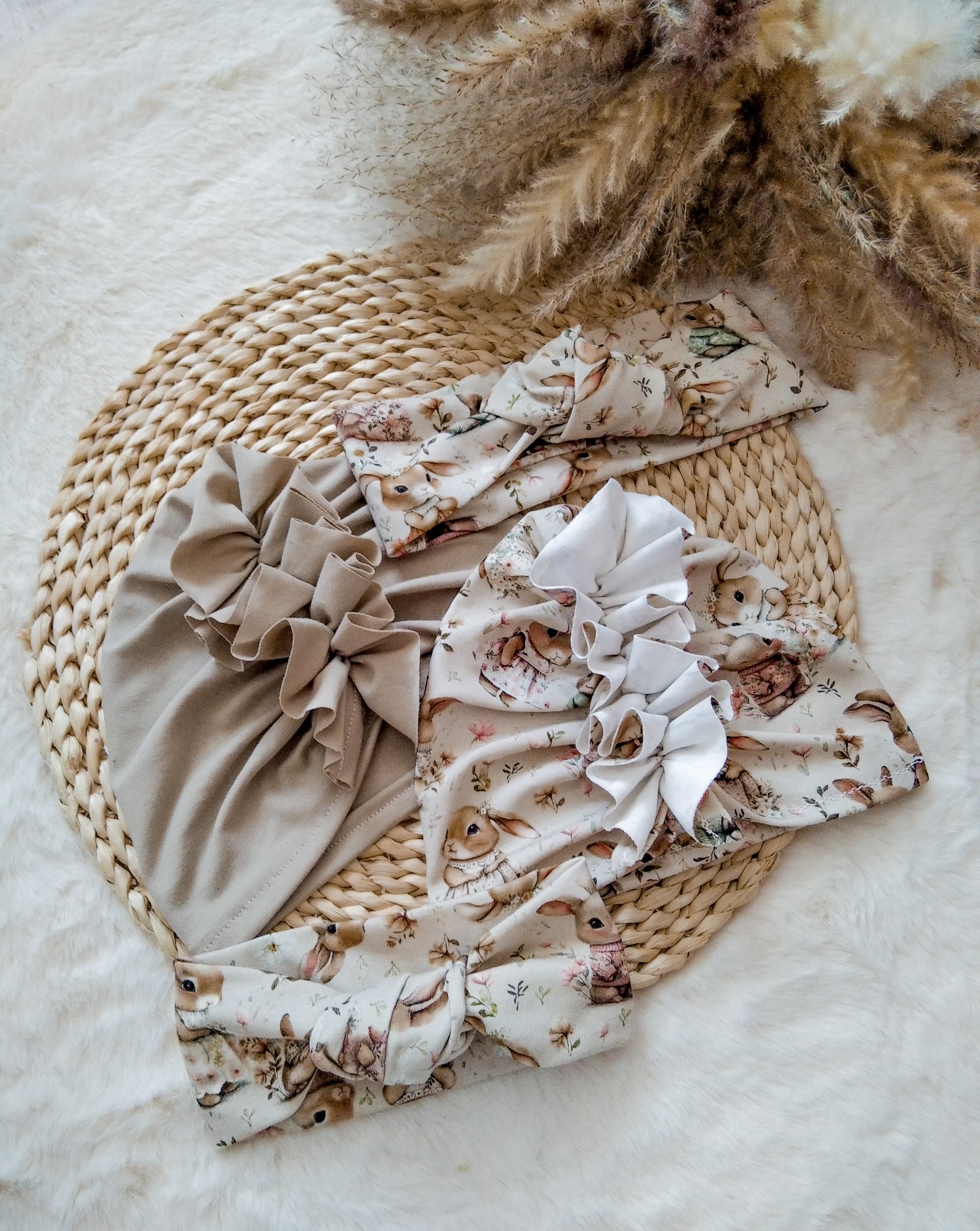 Beżowy turban z falbanką i opaski z motywem królików na słomianej podkładce, otoczone puszystym, białym futerkiem i suszonymi trawami w tle.