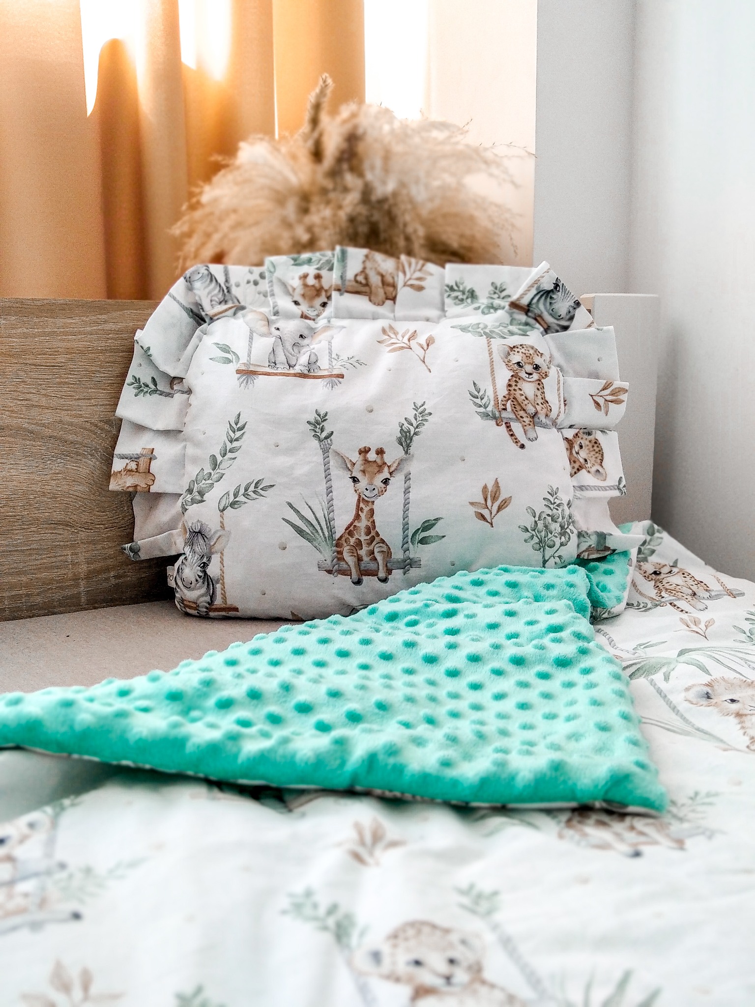 Dziecięca poduszka z falbanką i kocyk z wypustkami, w pastelowych barwach i motywem zwierząt na huśtawkach, leżące na pościeli z tym samym wzorem.