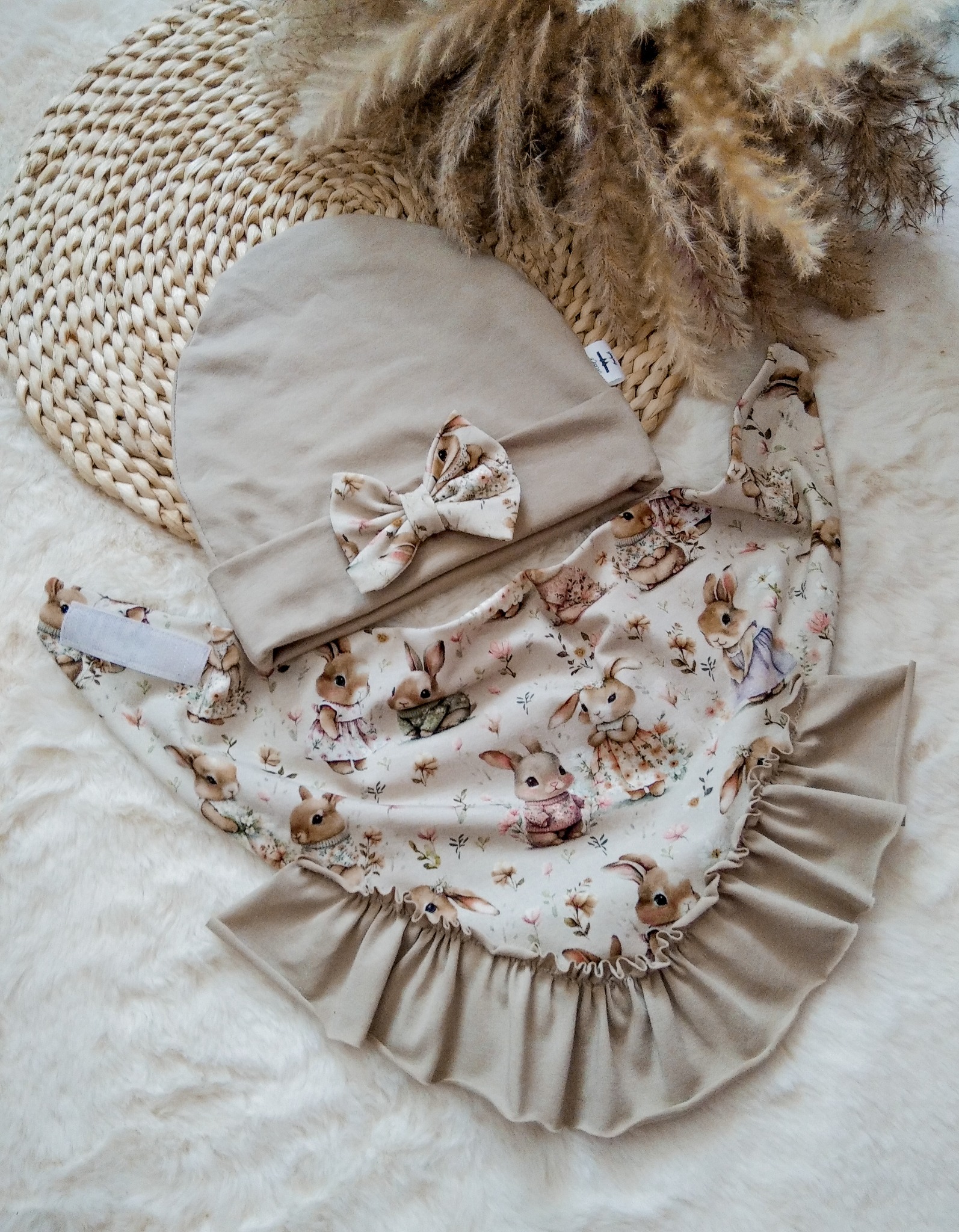 Beżowy komplet niemowlęcy: czapeczka z kokardką i śliniak z falbanką, w króliczki, na puszystym tle z dekoracjami. Uroczy zestaw dla dziecka, idealny na prezent.