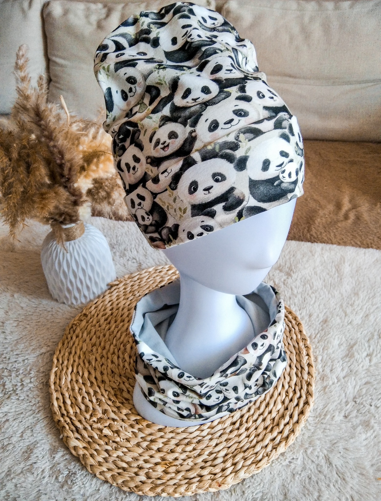 Ręcznie uszyty komplet: czapka i komin z motywem pand na manekinie, na słomianej podkładce. W tle wazon z suszonymi trawami i beżowa kanapa.