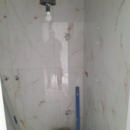 Lagrance - Wykończona ściana w łazience z marmurowymi płytkami, widoczny poziomica i wiadro z zaprawą. W tle odbicie osoby w oknie.