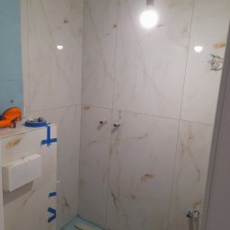 Lagrance - Wykończona ściana łazienki z marmurowymi płytkami, widoczne przyłącza wodne i narzędzia glazurnicze. Podłoga zabezpieczona folią. Oświetlenie sufitowe.