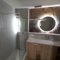 Lagrance - Nowoczesna łazienka z kabiną prysznicową typu walk-in, umywalką w szafce z drewnianym fornirem i okrągłym lustrem z podświetleniem LED na dekoracyjnej ścianie.