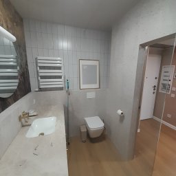 Lagrance - Jasna łazienka po remoncie: biała umywalka z złotą armaturą, toaleta, grzejnik drabinkowy i lustro z drewnianym akcentem. Minimalistyczny design z drewnianą podłogą.