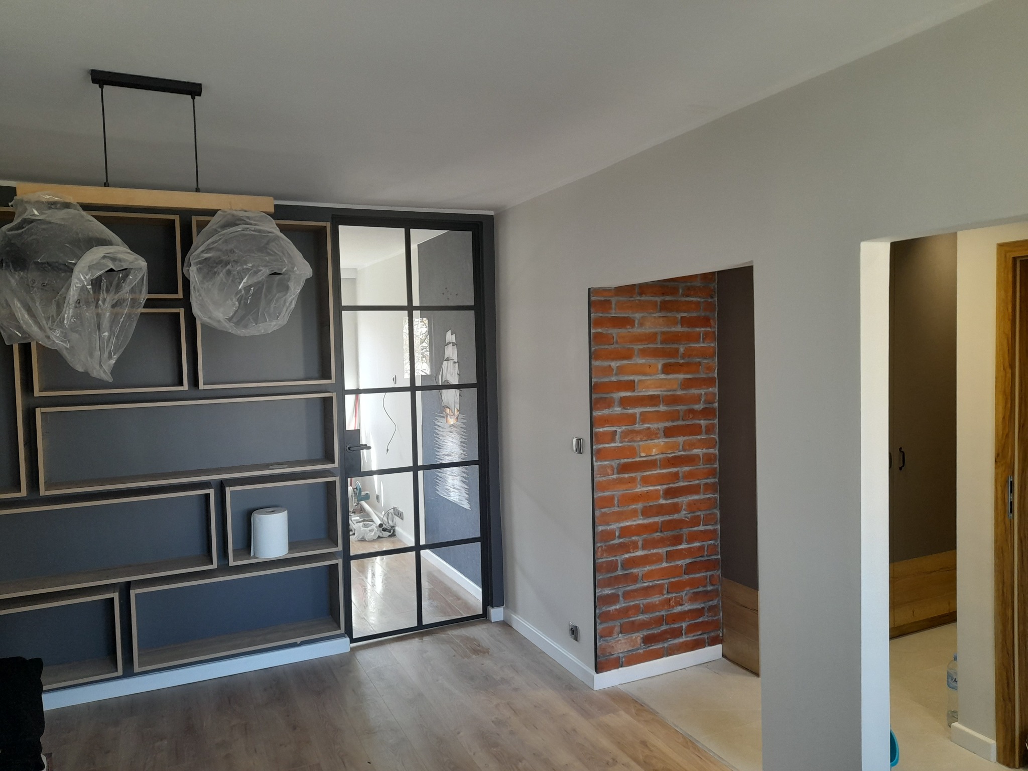 Nowoczesne wnętrze z drewnianą podłogą, szarą ścianą z półkami, przeszklonymi drzwiami i fragmentem ściany z czerwonej cegły. Lampy wiszące zabezpieczone folią.