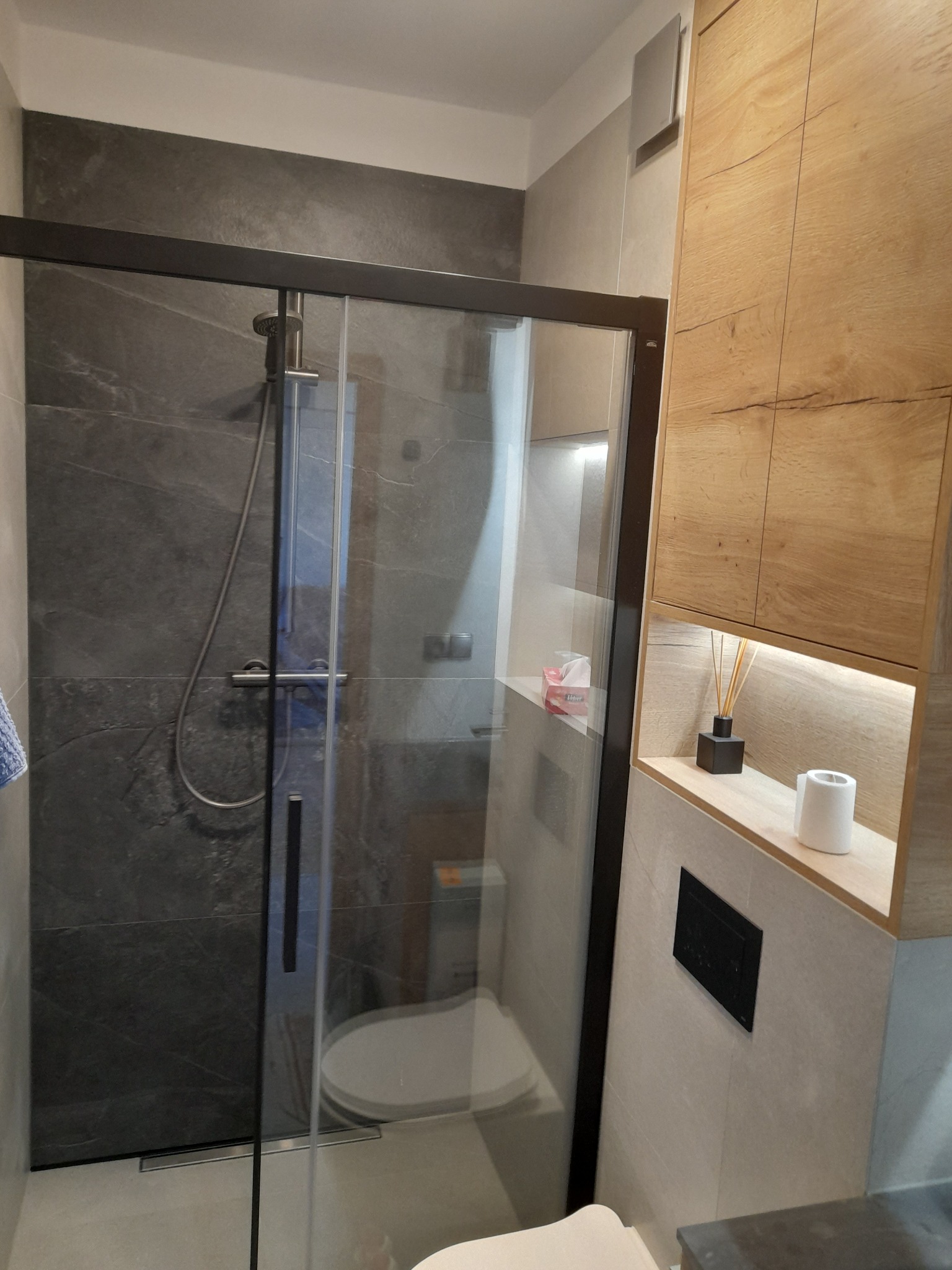 Nowoczesna kabina prysznicowa z czarną ramą i szarymi płytkami imitującymi kamień. Drewniana szafka z oświetleniem LED i dekoracjami. Minimalistyczny design łazienki.