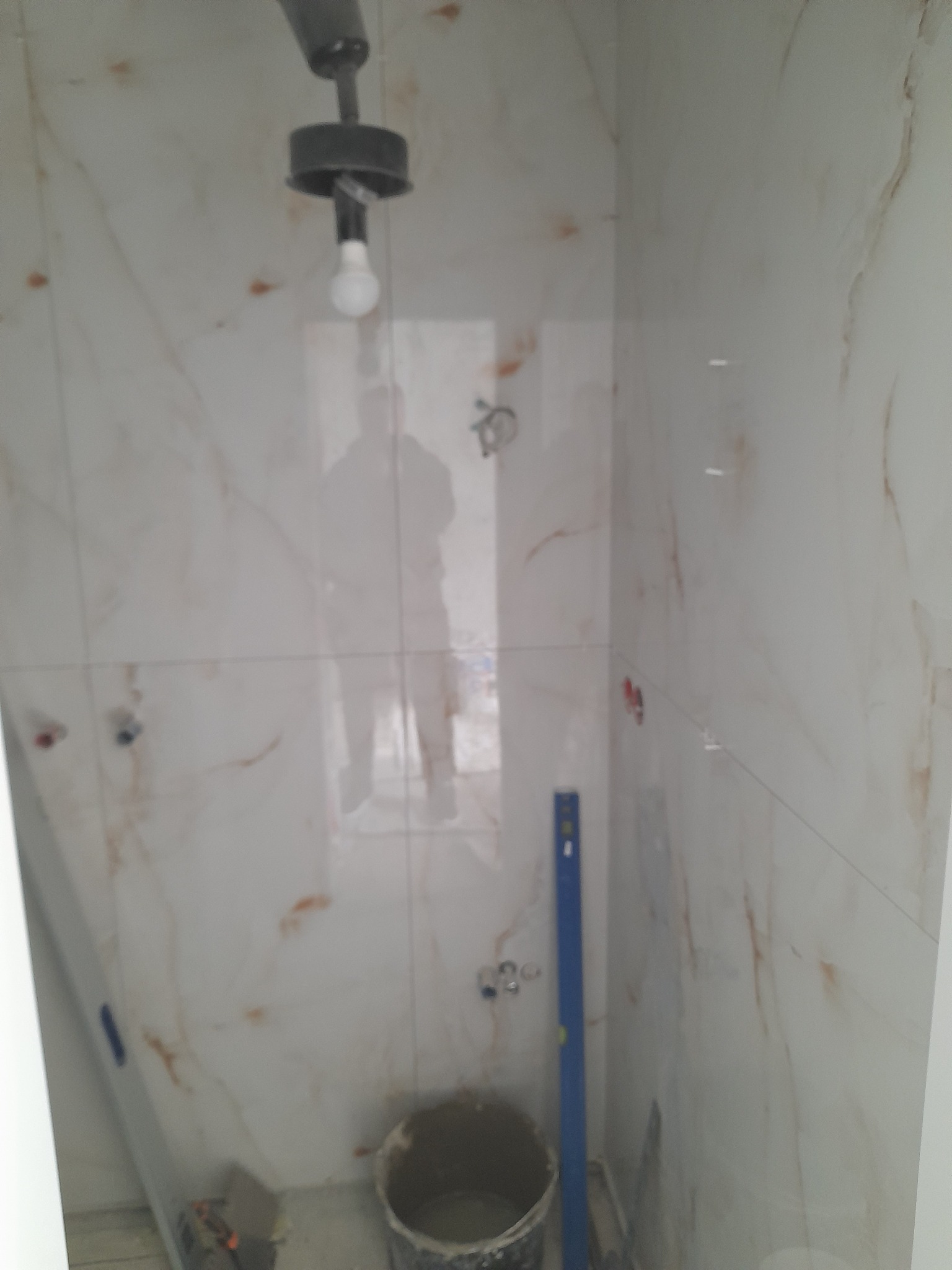 Wykończona ściana w łazience z marmurowymi płytkami, widoczny poziomica i wiadro z zaprawą. W tle odbicie osoby w oknie.