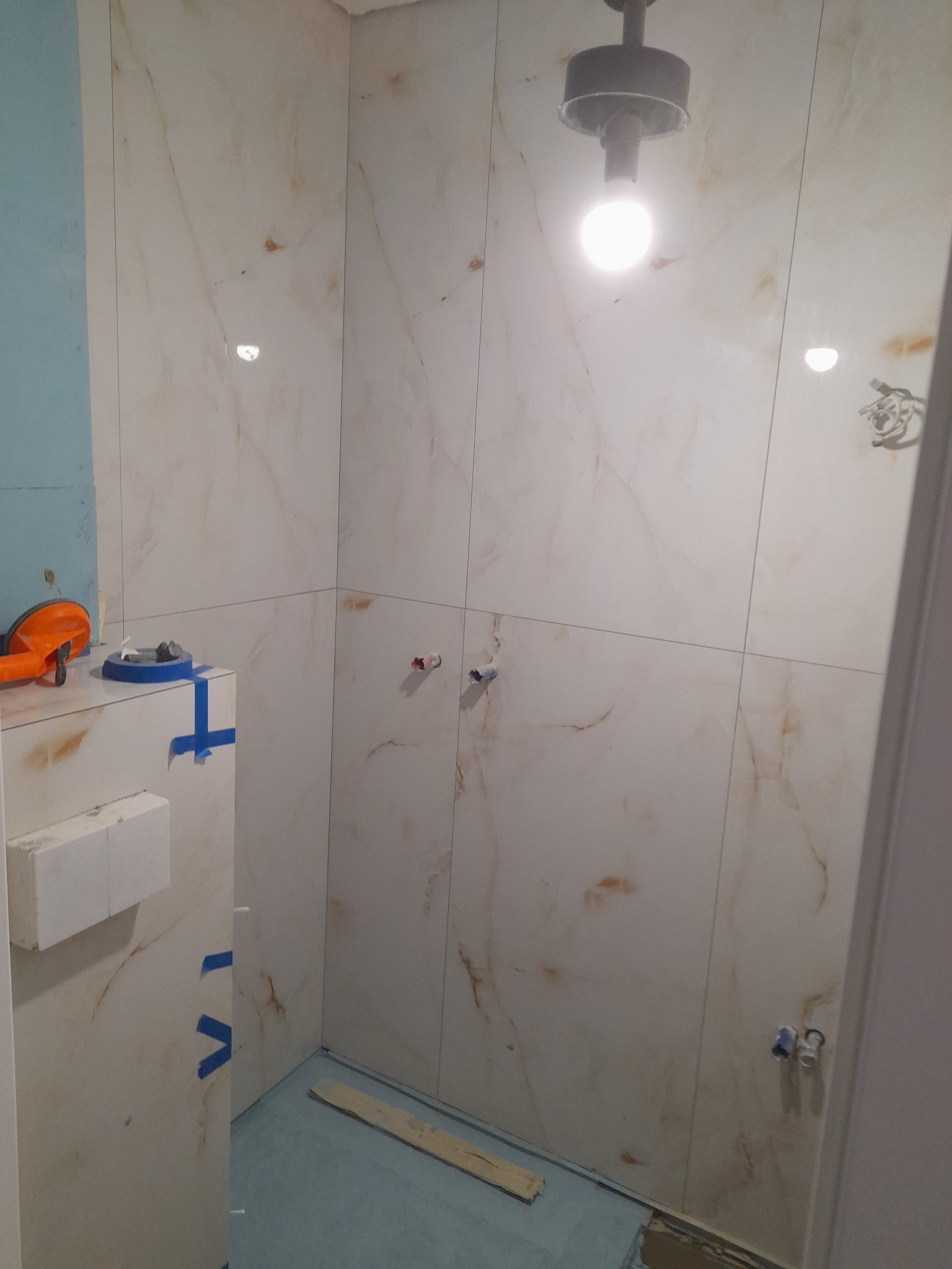 Wykończona ściana łazienki z marmurowymi płytkami, widoczne przyłącza wodne i narzędzia glazurnicze. Podłoga zabezpieczona folią. Oświetlenie sufitowe.