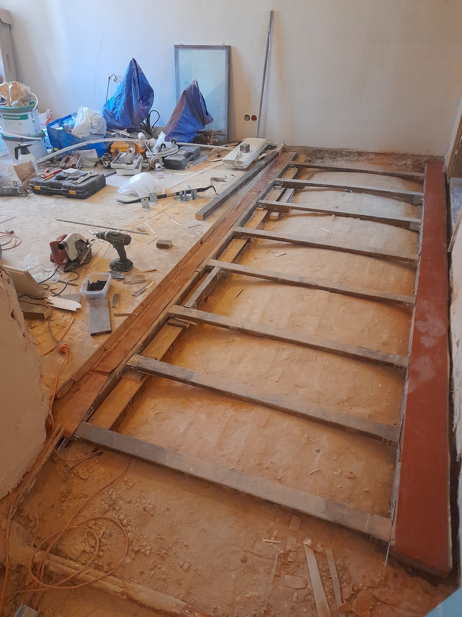 Drewniana konstrukcja pod podłogę w trakcie montażu, widoczne narzędzia i materiały budowlane, surowy stan wnętrza, przewody elektryczne na podłodze.